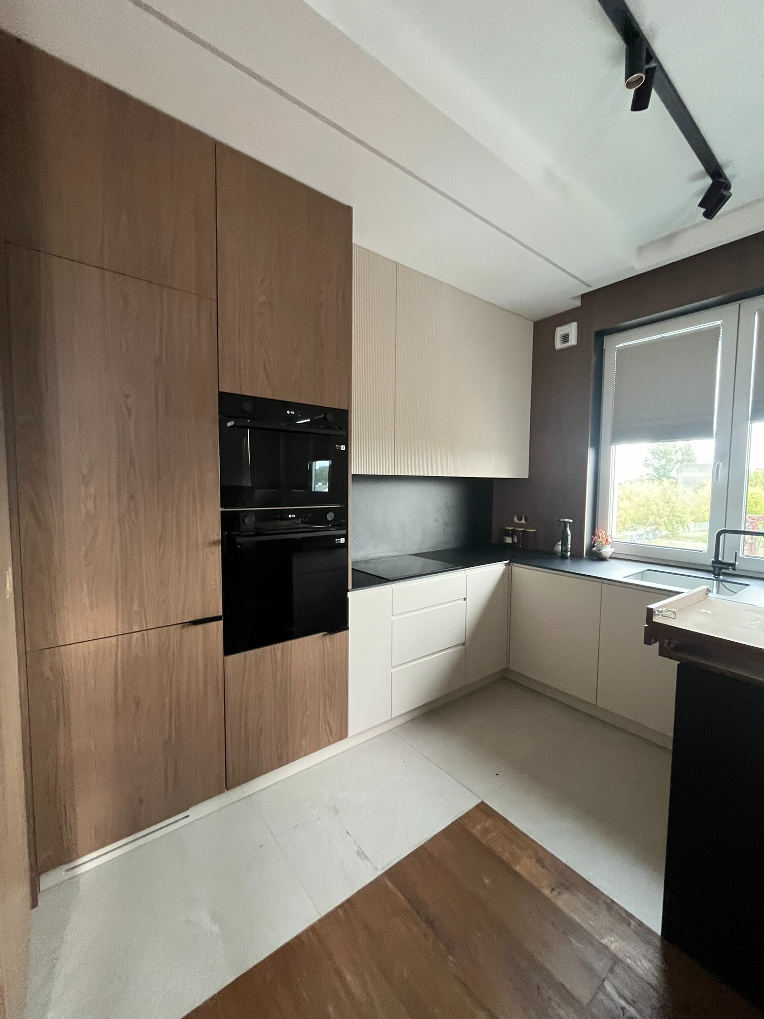 Nowoczesna kuchnia z frontami w odcieniach drewna i beżu, czarnym blatem i oświetleniem szynowym. Widok na okno z roletą i fragment drewnianego stołu.