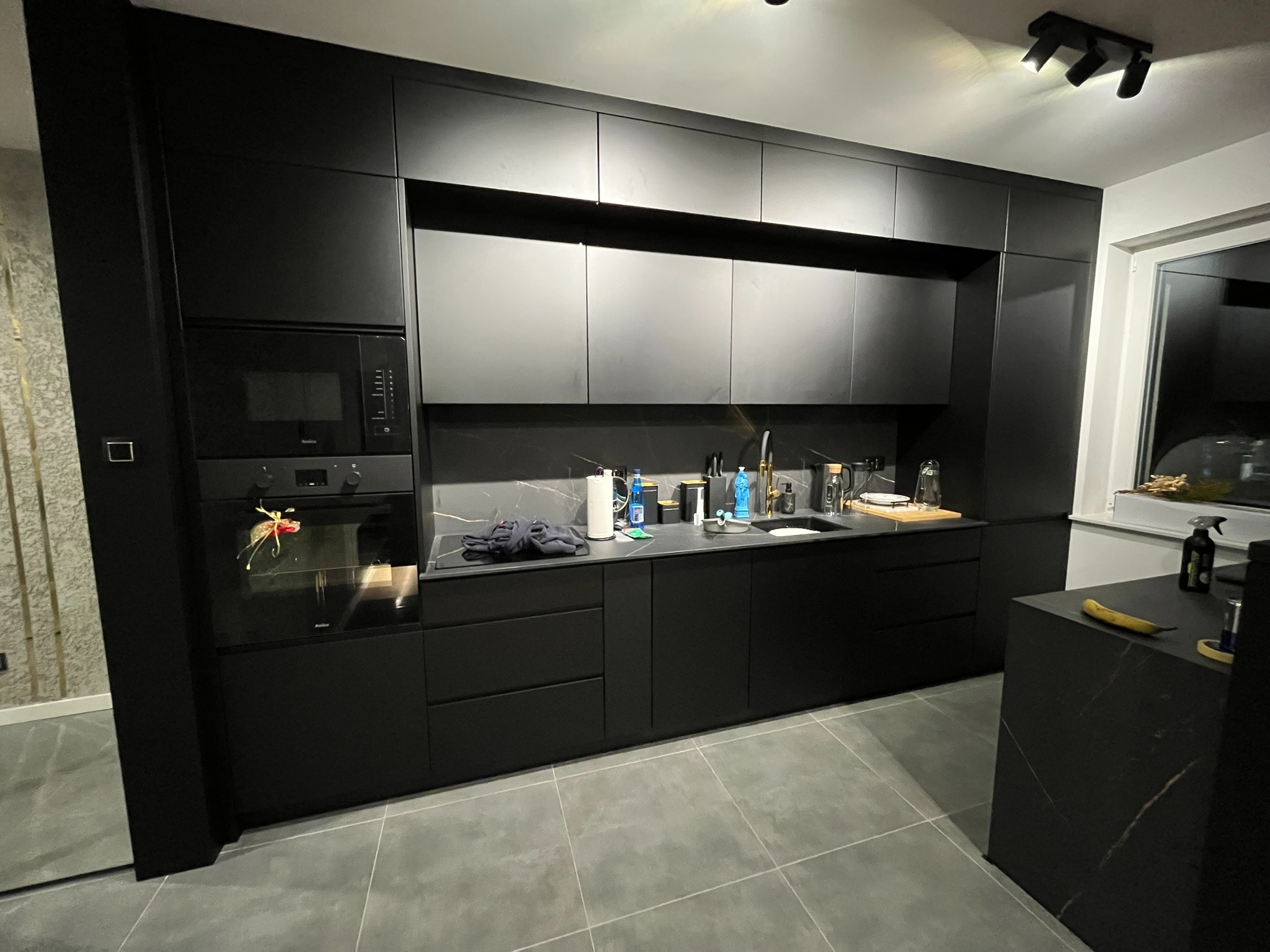 Nowoczesna, czarna kuchnia na wymiar z marmurowym blatem i wyspą. Minimalistyczny design, oświetlenie punktowe i sprzęt AGD w zabudowie.