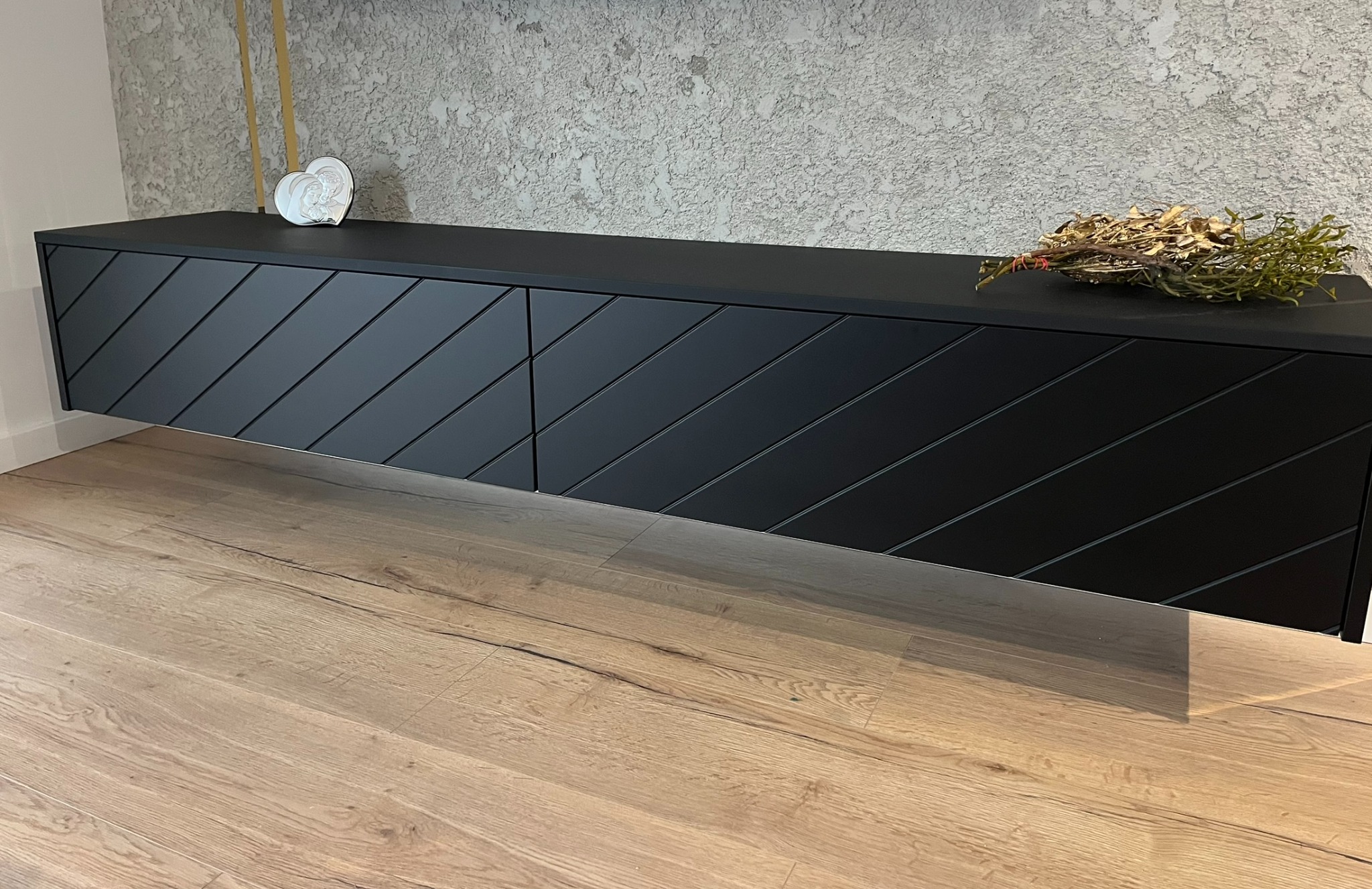 Czarna, wisząca szafka RTV z ukośnymi żłobieniami na frontach, dekoracje na blacie, na tle ściany o fakturze betonu i drewnianej podłogi.