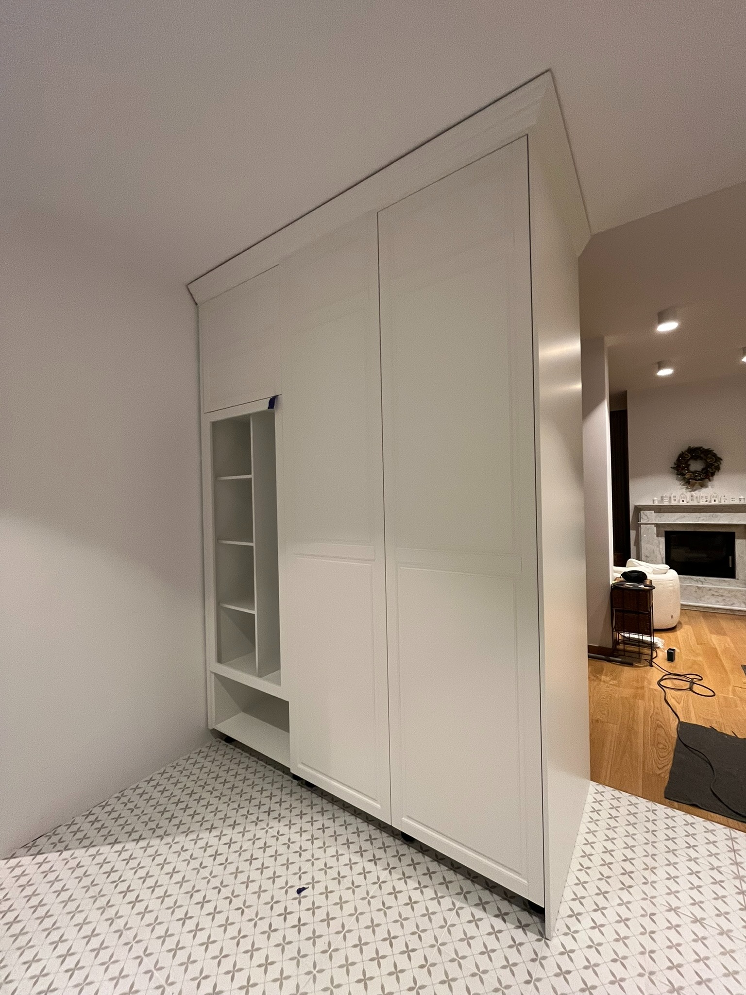 Biała szafa wnękowa z otwartymi półkami w nowoczesnym wnętrzu. Widoczny fragment salonu z kominkiem. Dekoracyjne płytki podłogowe.