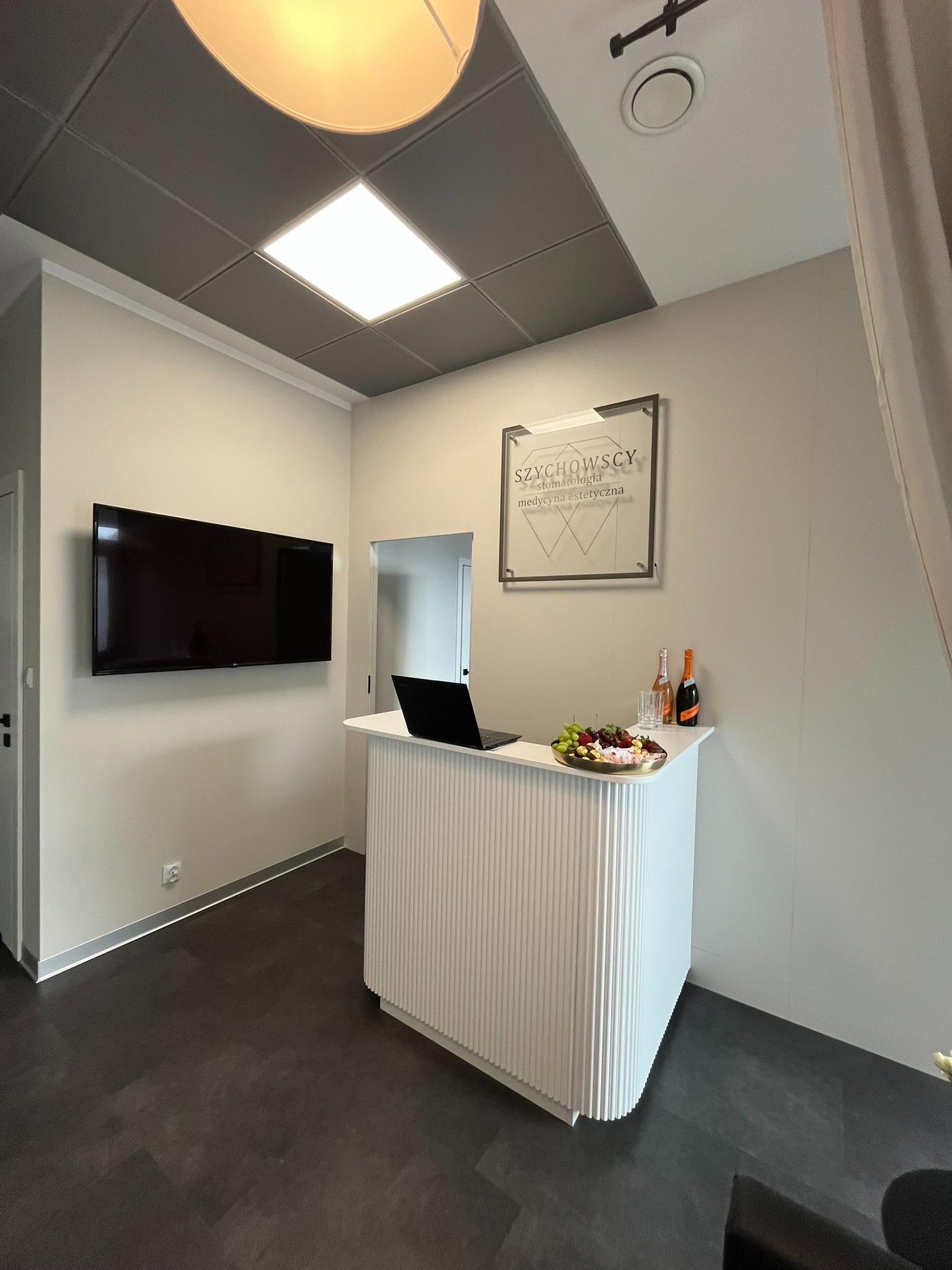 Nowoczesna recepcja z białym, żłobkowanym blatem, laptopem i poczęstunkiem. W tle logo firmy na ścianie i telewizor. Minimalistyczny design wnętrza.
