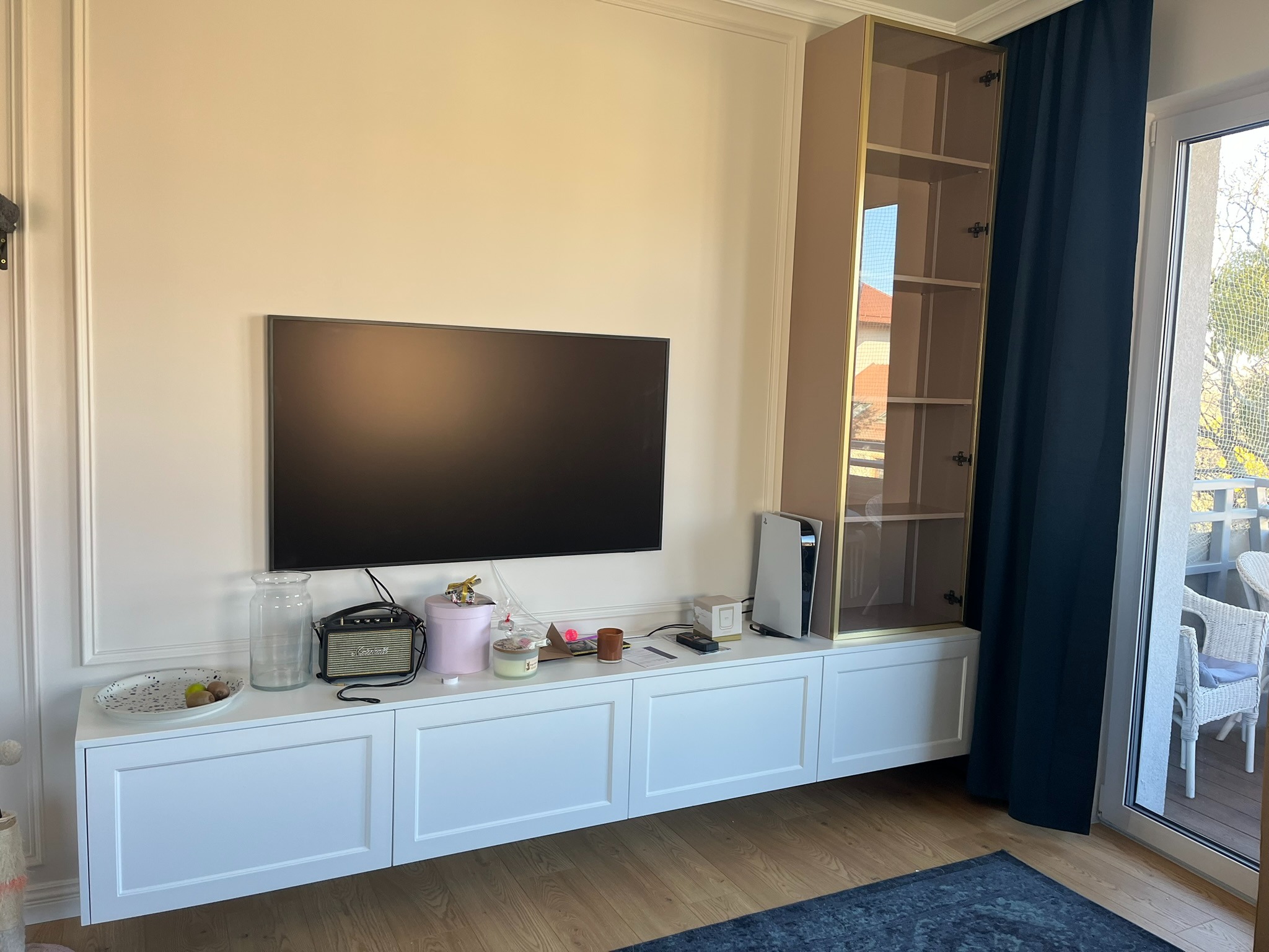 Biała, niska szafka RTV z dekoracyjnymi frontami i witryną z brązowymi ramami, na tle ściany z listwami. Na szafce akcesoria i konsola do gier.