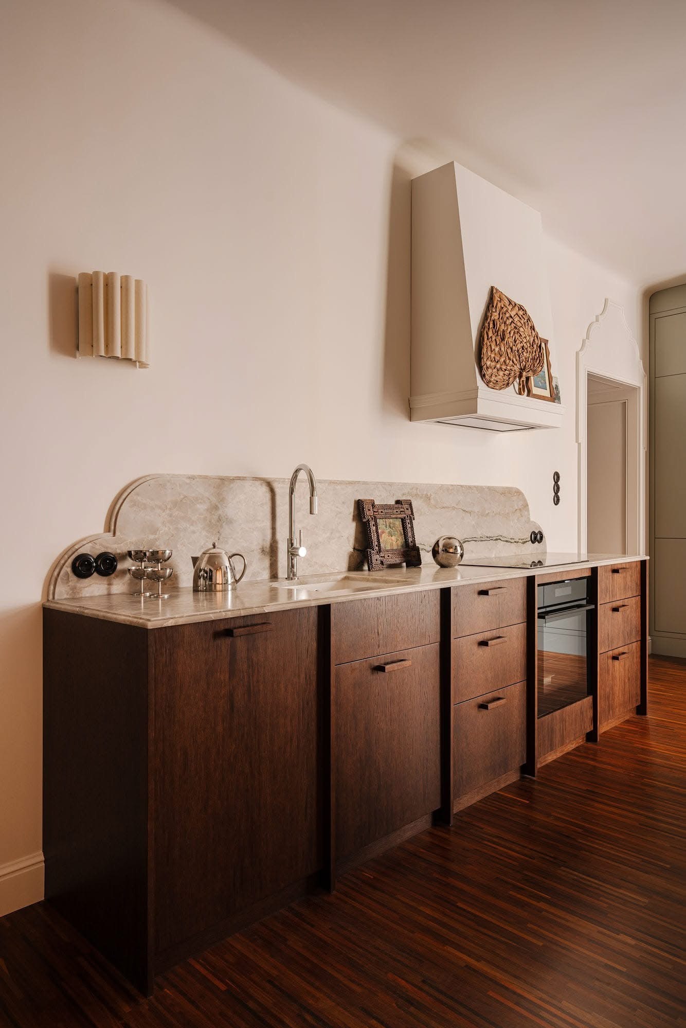 Kuchnia z ciemnego drewna i marmurowym blatem, zintegrowana płyta grzewcza i piekarnik. Dekoracje: lampa ścienna, plecionka, ramka. Parkiet w jodełkę.