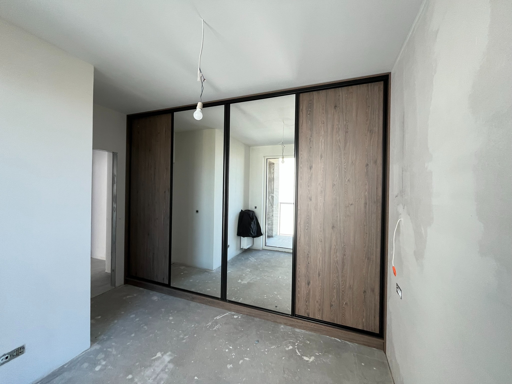 Szafa wnękowa z przesuwnymi drzwiami lustrzanymi i panelami drewnopodobnymi w surowym wnętrzu. Widoczne niedokończone ściany i podłoga.