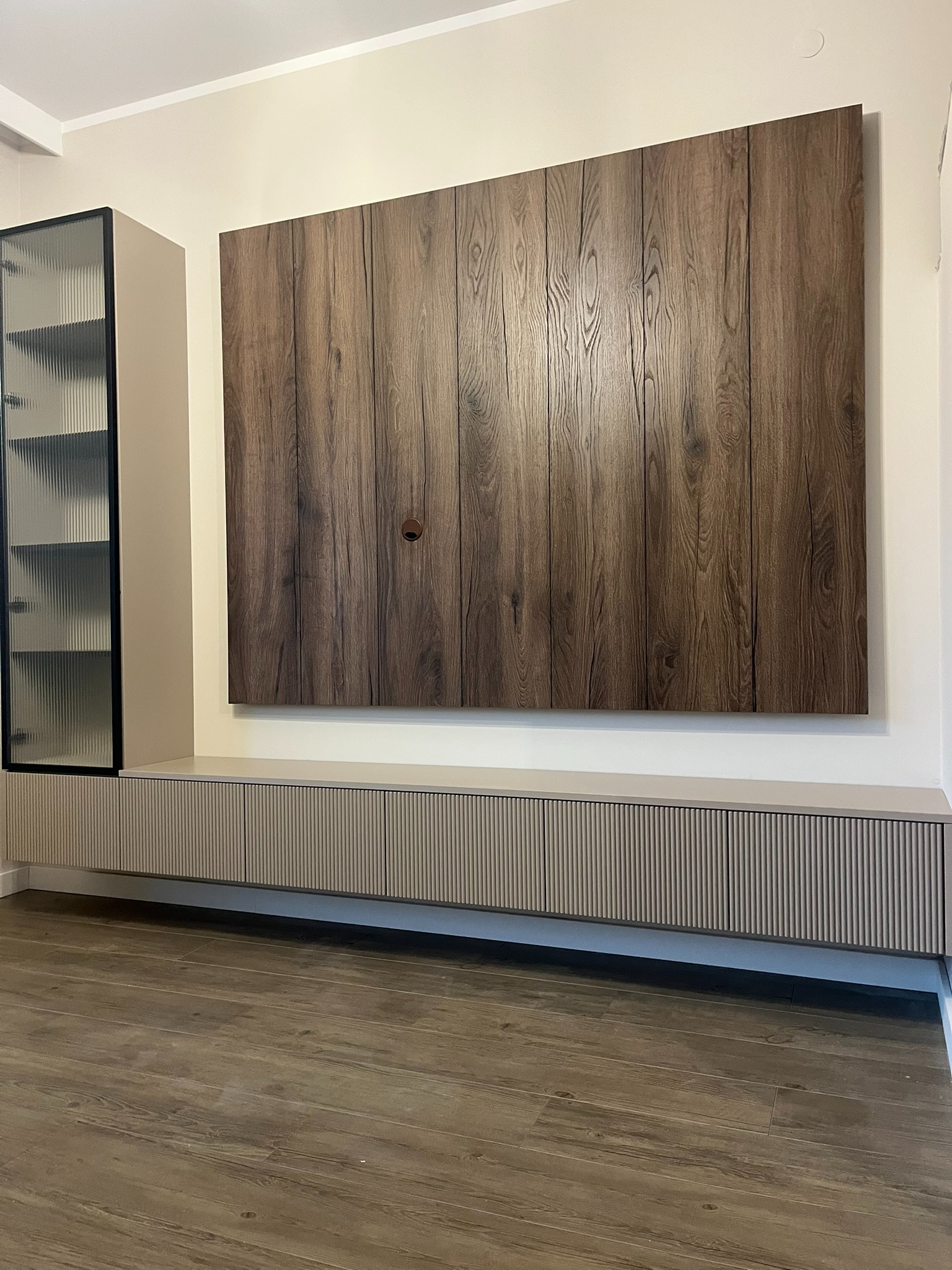 Nowoczesna szafka RTV z drewnianym panelem ściennym i szklaną witryną. Ryflowane fronty szafek i podłoga imitująca drewno tworzą spójną całość.