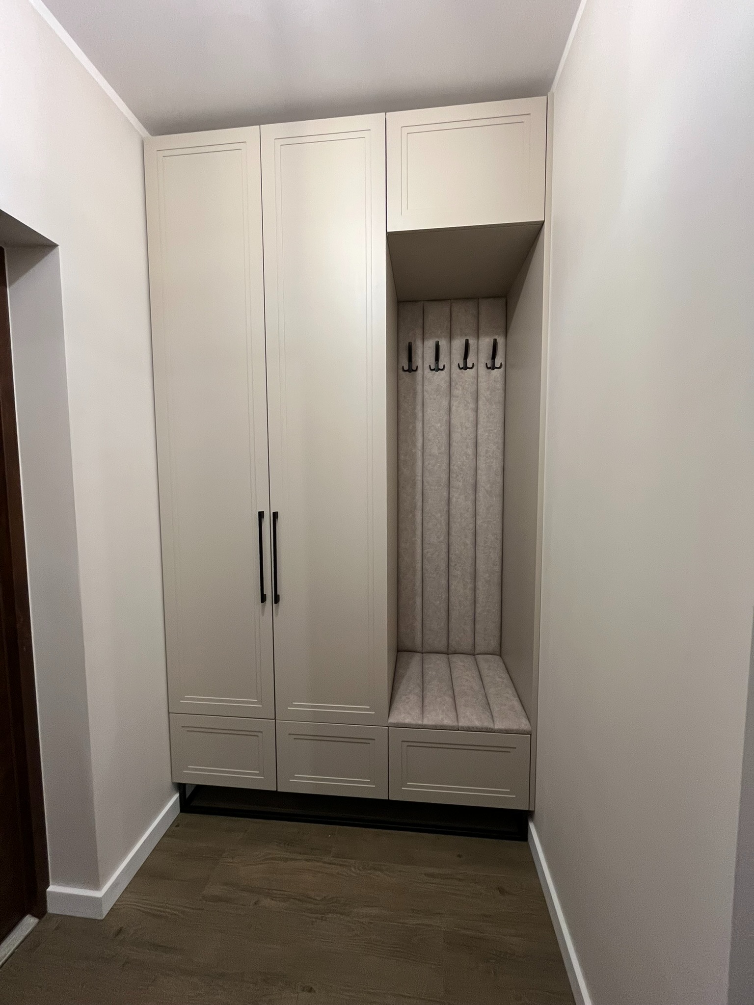 Jasna zabudowa przedpokoju z szafą i siedziskiem z wieszakami, w tonacji beżowej. Minimalistyczne czarne uchwyty i subtelne detale wykończeniowe.