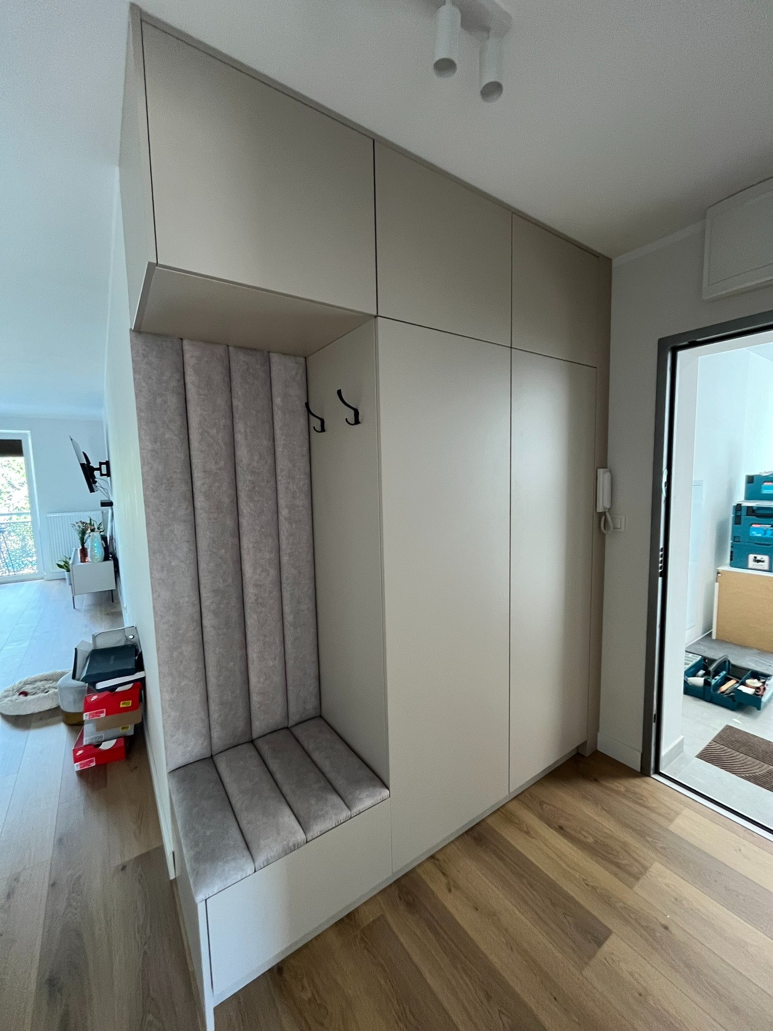 Jasna zabudowa przedpokoju z siedziskiem i wieszakami, minimalistyczny design, drewniana podłoga, widok na wnętrze mieszkania przez otwarte drzwi.