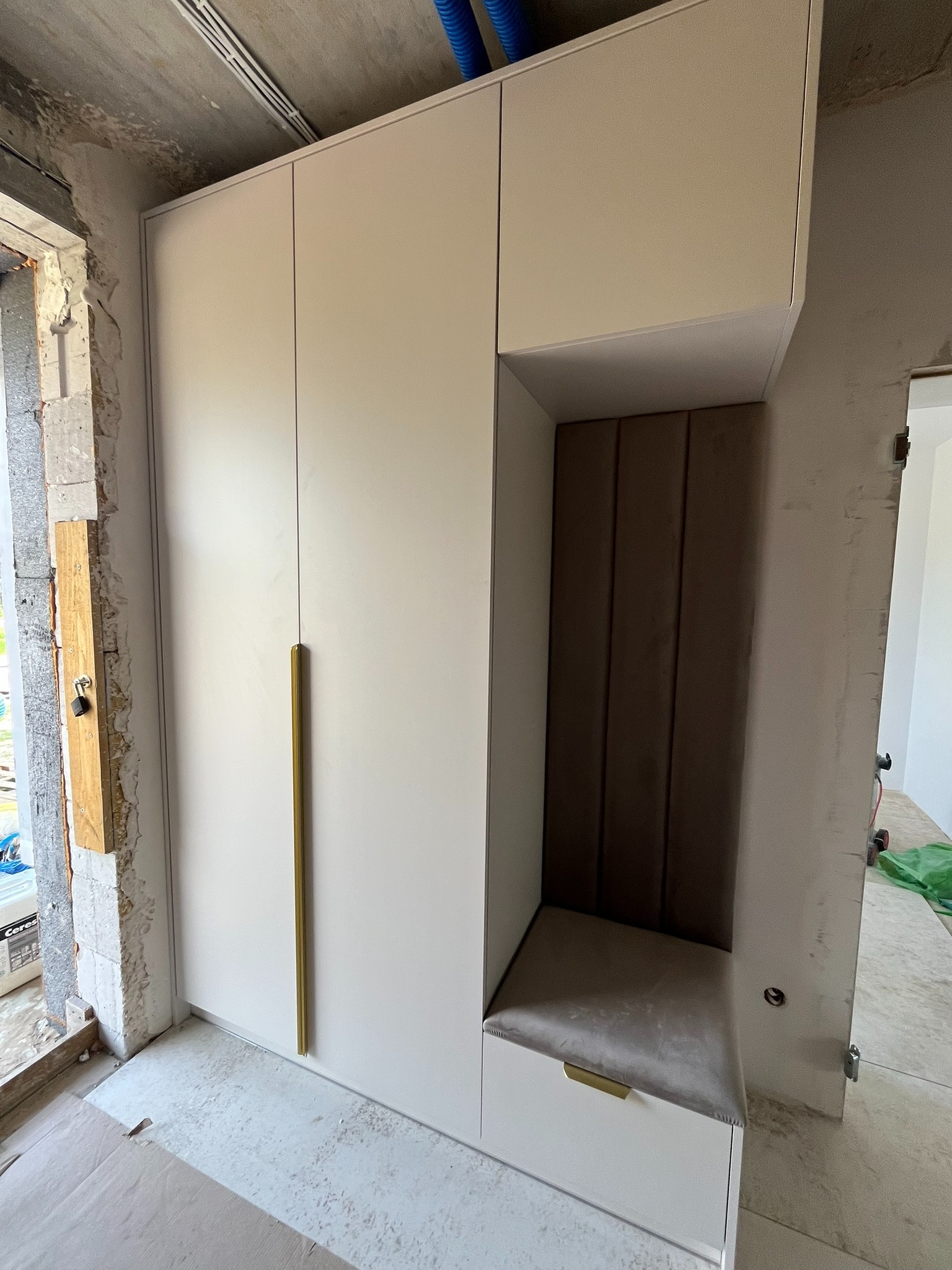 Biała szafa z siedziskiem i złotymi uchwytami na tle surowego wnętrza w trakcie wykończenia, widoczne elementy instalacji i materiały budowlane.