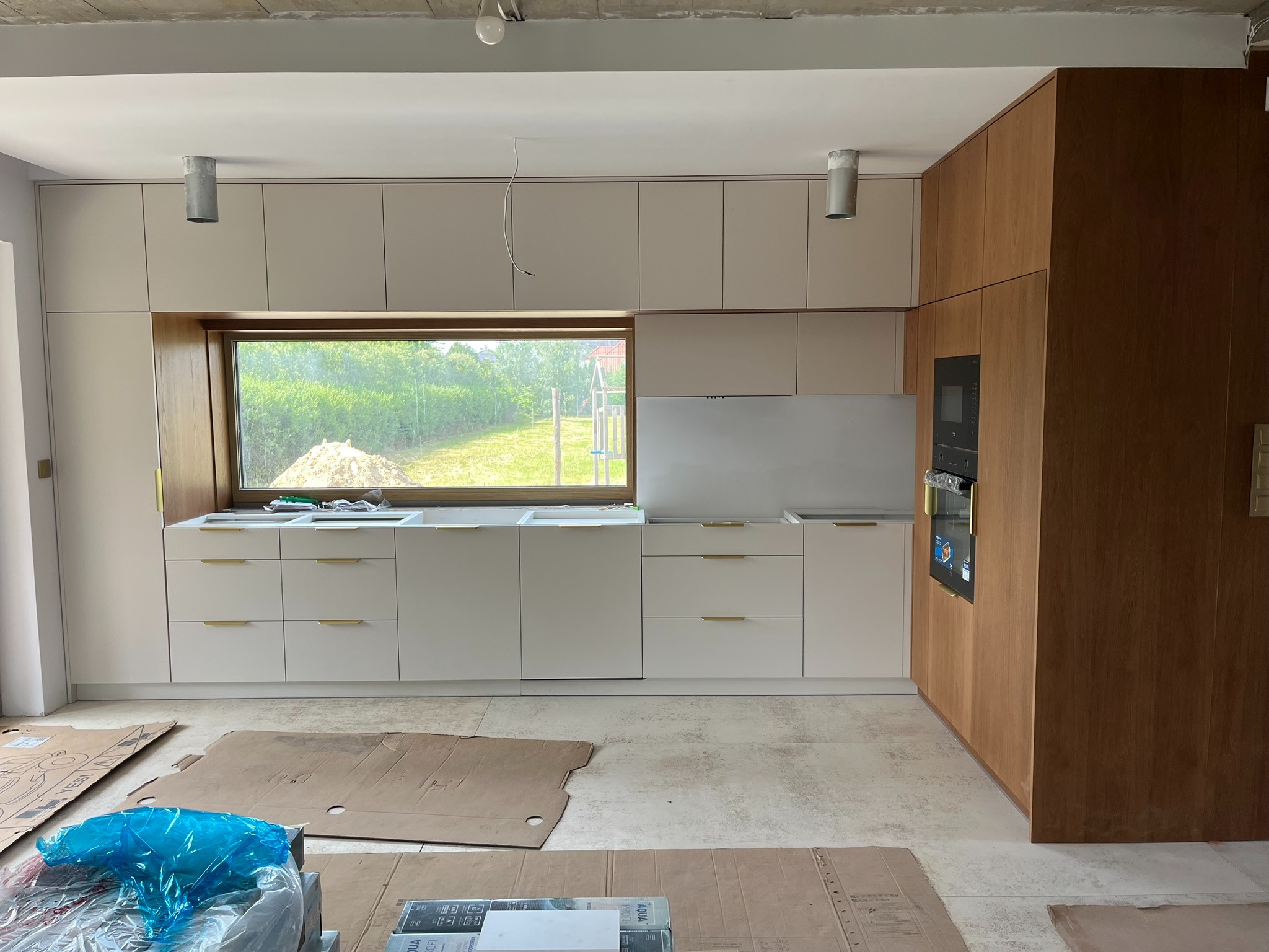 Jasna kuchnia z drewnianymi akcentami w trakcie montażu. Widoczne kartony ochronne na podłodze, okno z widokiem na zieleń. Uwagę zwracają złote uchwyty szafek.