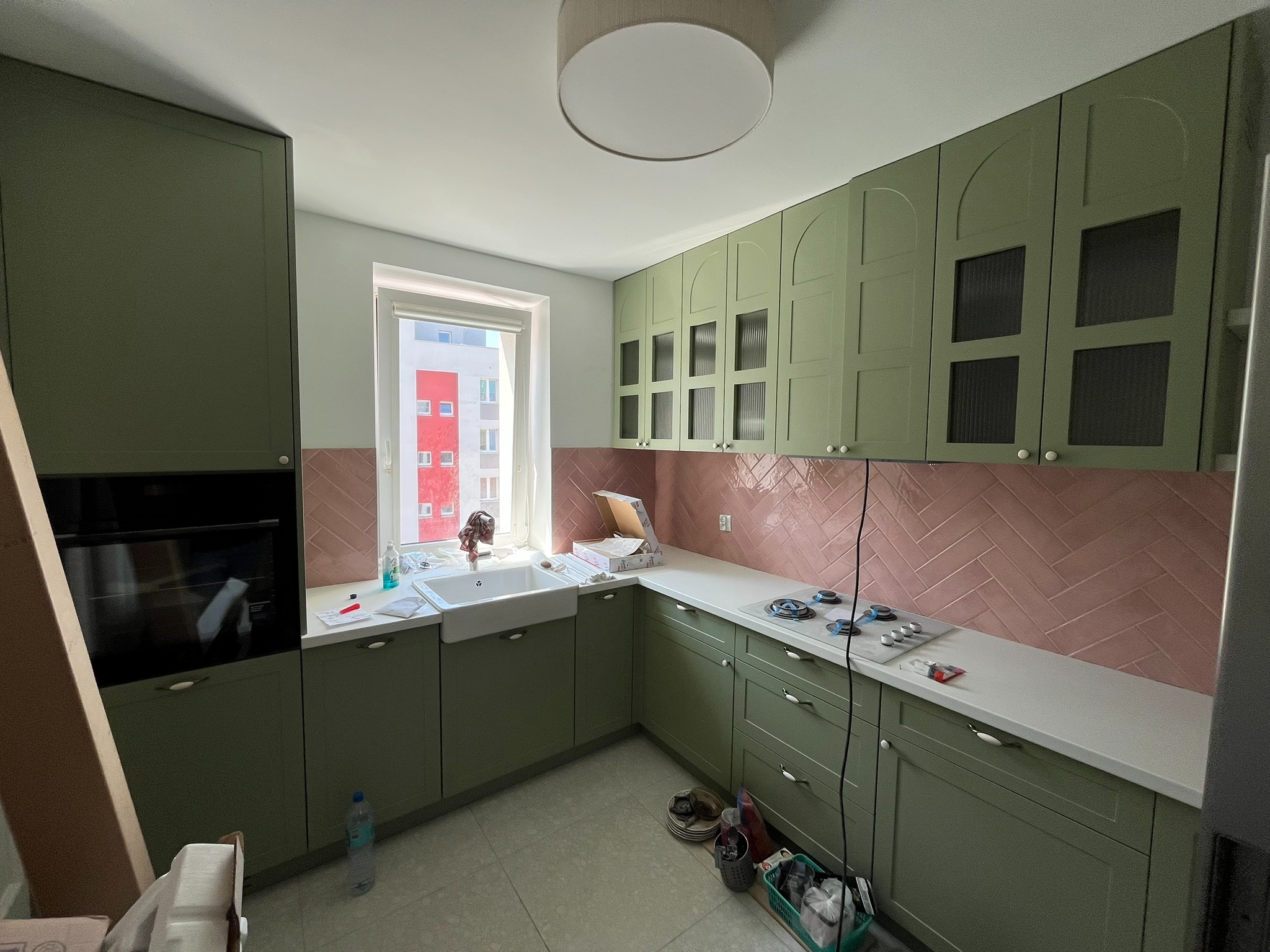 Kuchnia w odcieniach zieleni z różowymi płytkami ułożonymi w jodełkę na ścianie. Widoczny zlew, okno i częściowo zamontowany piekarnik.