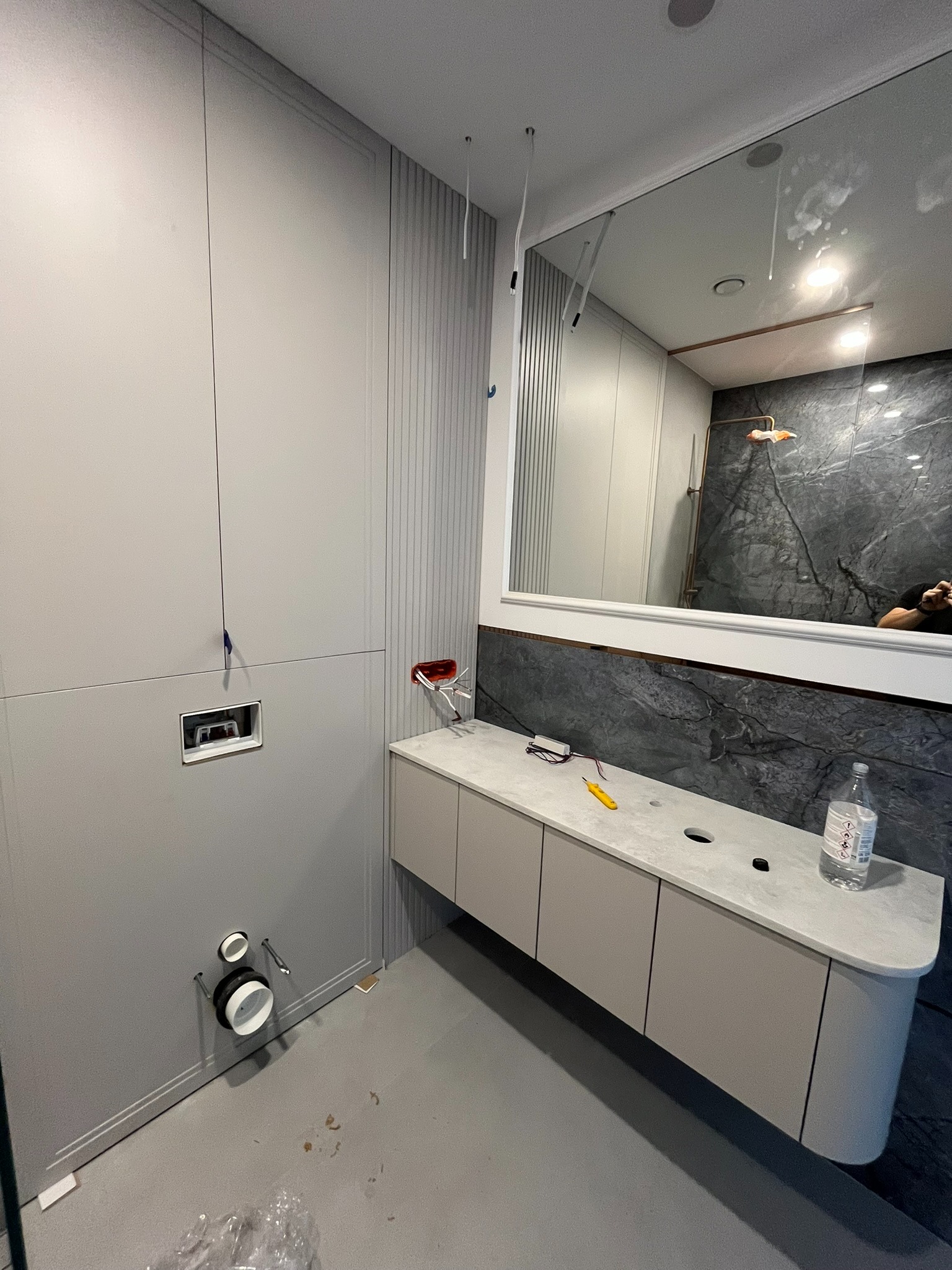 Szara zabudowa łazienkowa z szafką pod umywalkę z blatem, widoczne przyłącza wodno-kanalizacyjne w ścianie. Elementy wykończeniowe w trakcie montażu.