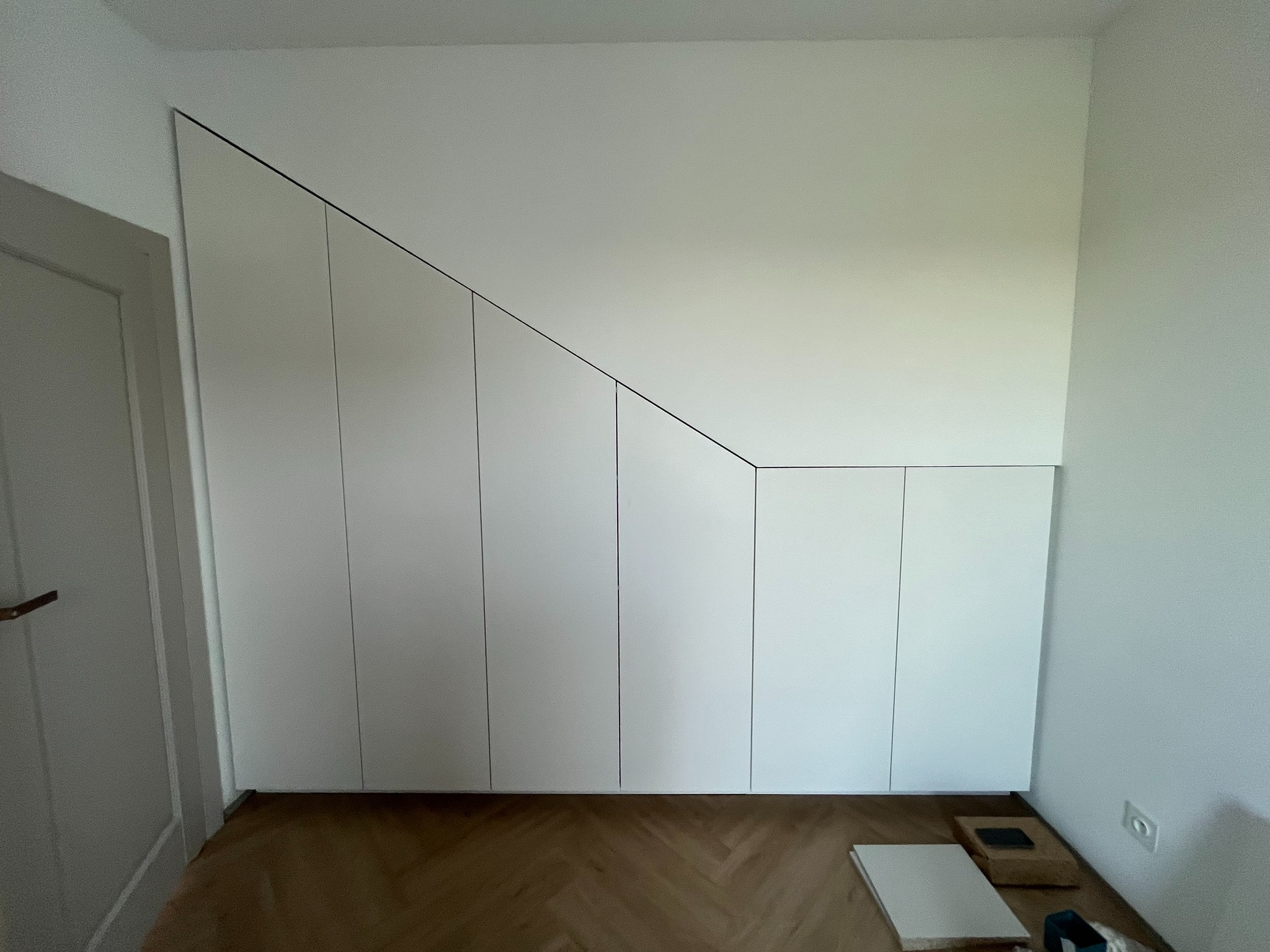 Biała szafa wnękowa na wymiar, dopasowana do skosu dachu, z widocznymi elementami montażowymi na parkiecie. Minimalistyczny design.