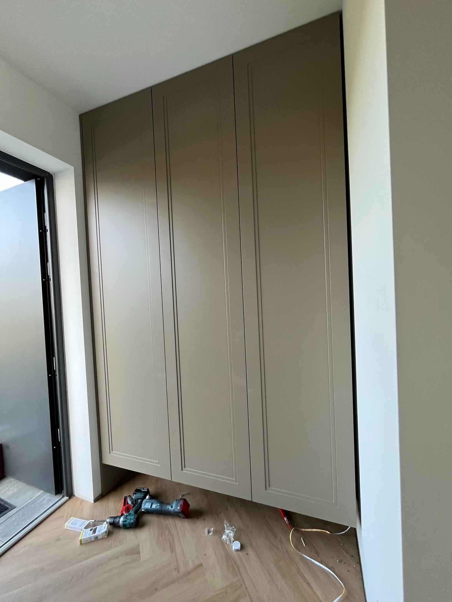 Jasnobrązowa szafa wnękowa z frezowanymi drzwiami, zamontowana w Strzałkowie, z narzędziami na podłodze. Widoczne listwy przypodłogowe i drzwi.