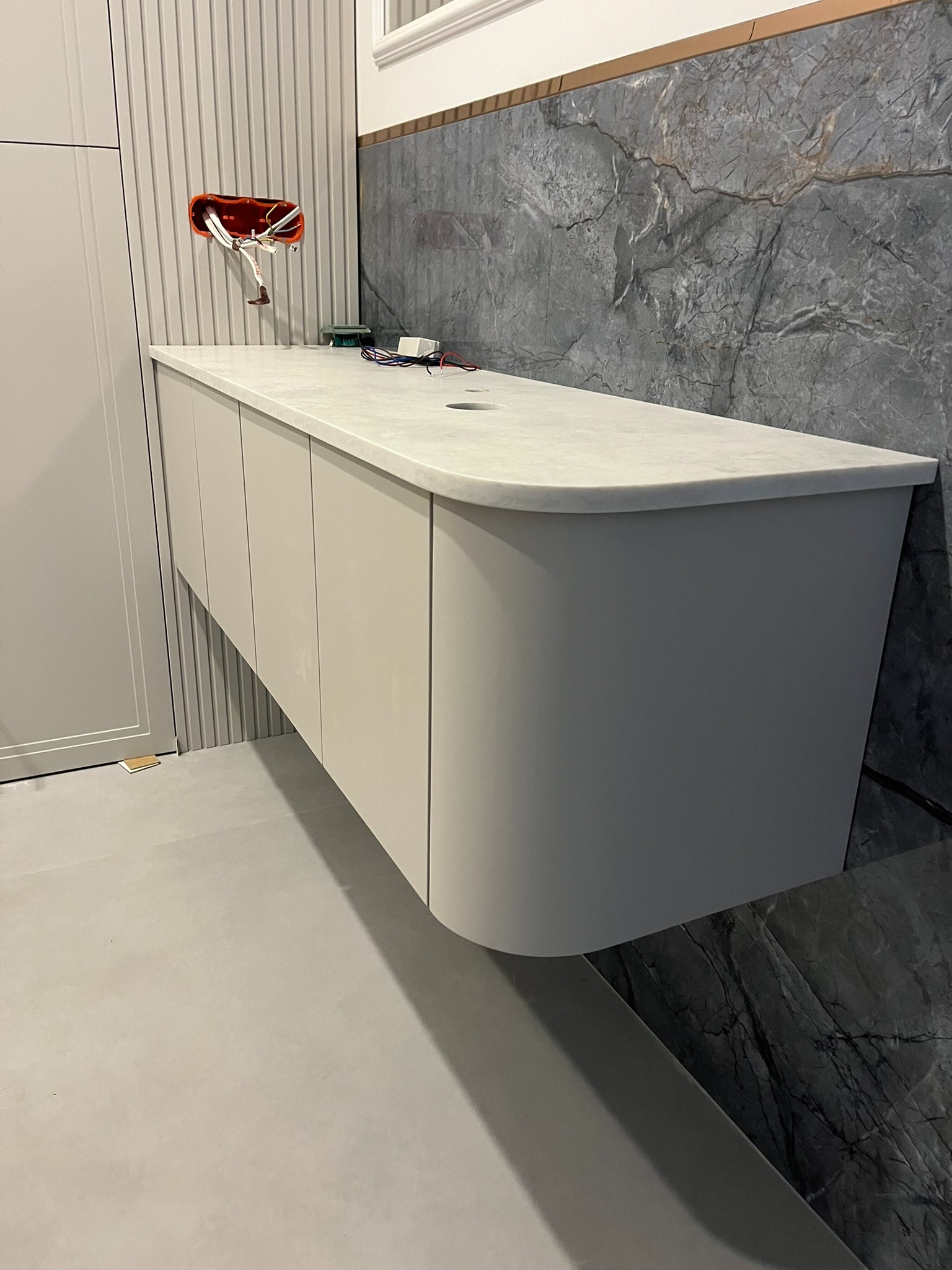 Beżowa szafka łazienkowa z zaokrąglonymi krawędziami, marmurowa ściana w tle, widoczne niedokończone podłączenia elektryczne, nowoczesny design.