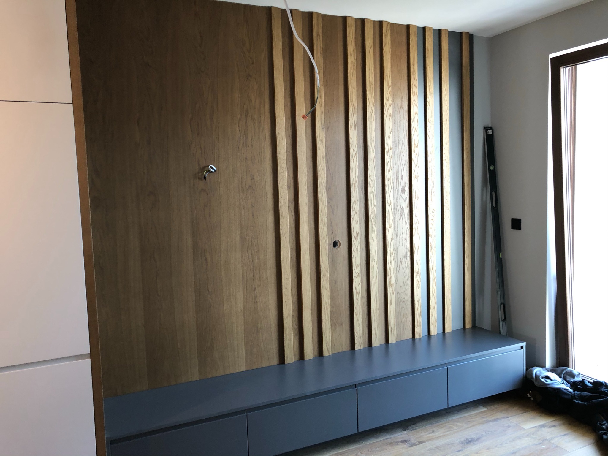 Nowoczesna zabudowa ściany z drewnianymi lamelami i szarą szafką RTV. Widoczne przewody i poziomica, sugerujące etap montażu. Jasna podłoga i ściany.