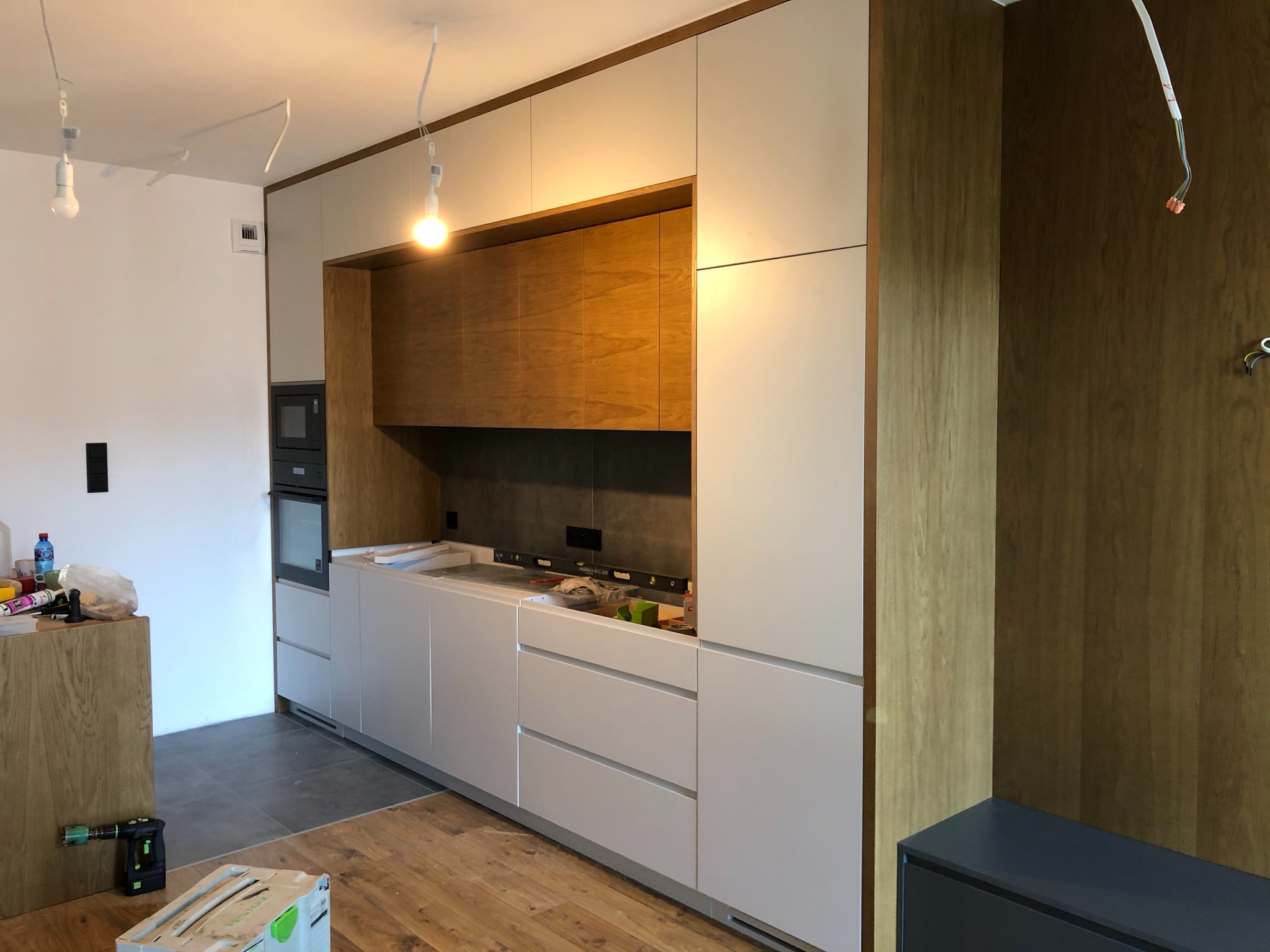 Montaż nowoczesnej kuchni: białe fronty, drewniane akcenty, surowy beton na ścianie. Widoczne narzędzia i poziomica, podkreślające etap wykończenia wnętrza.