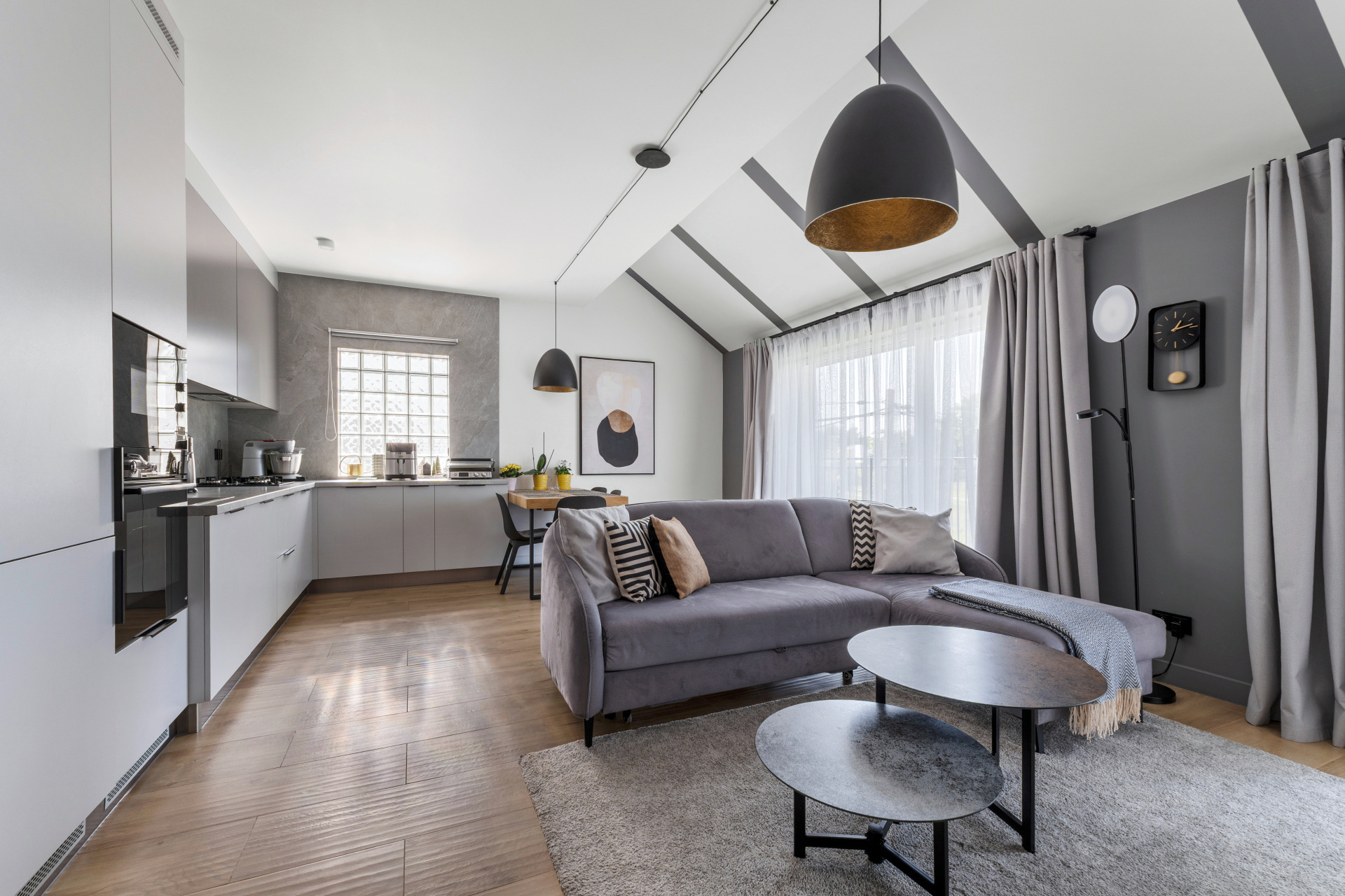 Stylowe wnętrze: szara sofa narożna z poduszkami, stoliki kawowe na dywanie, kuchnia z szarymi szafkami, okno z luksferami, lampy wiszące i skośny sufit.