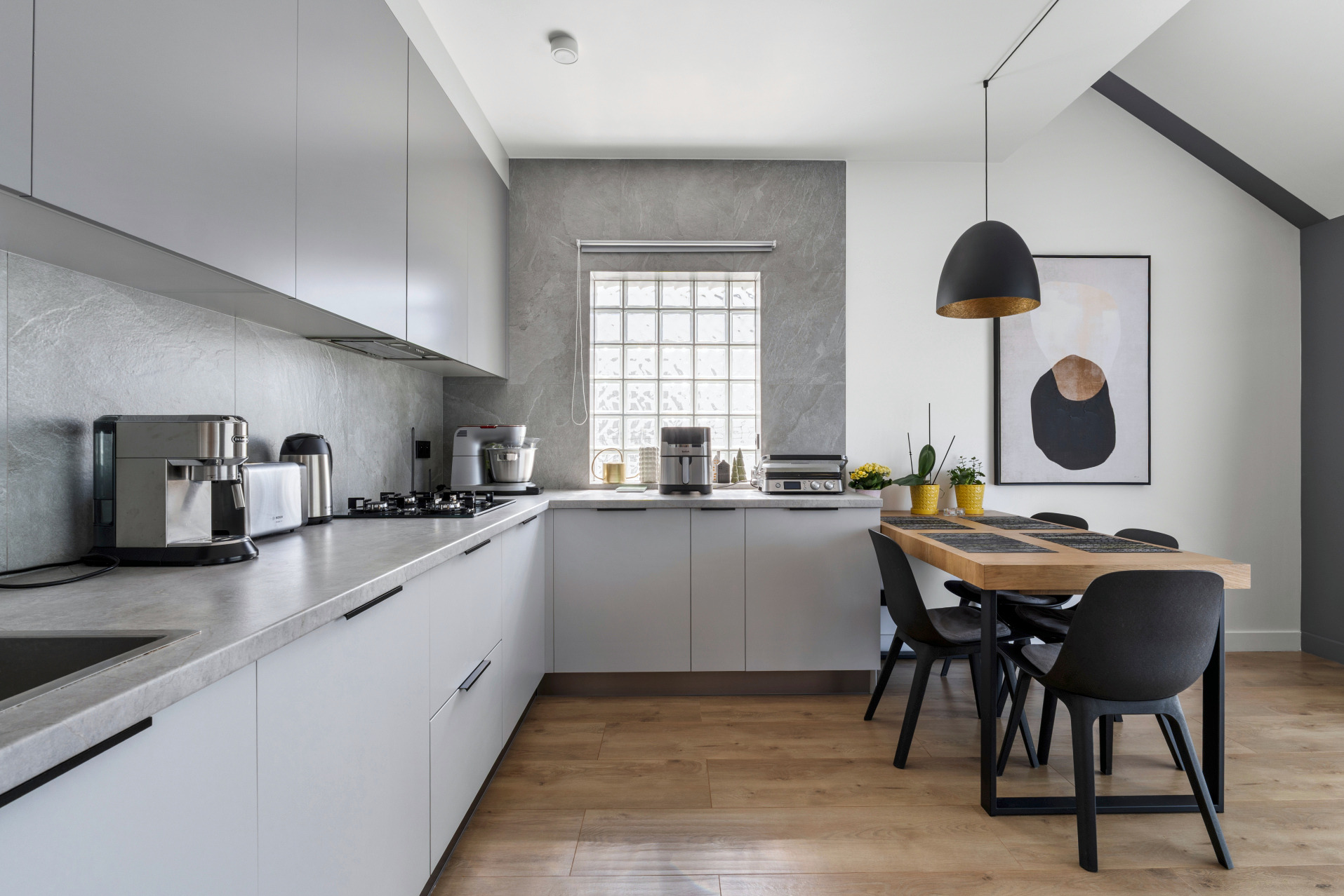 Nowoczesna kuchnia z szarymi frontami, drewnianym stołem i czarnymi krzesłami. Na blacie ekspres do kawy i inne sprzęty. Okno z luksferów.