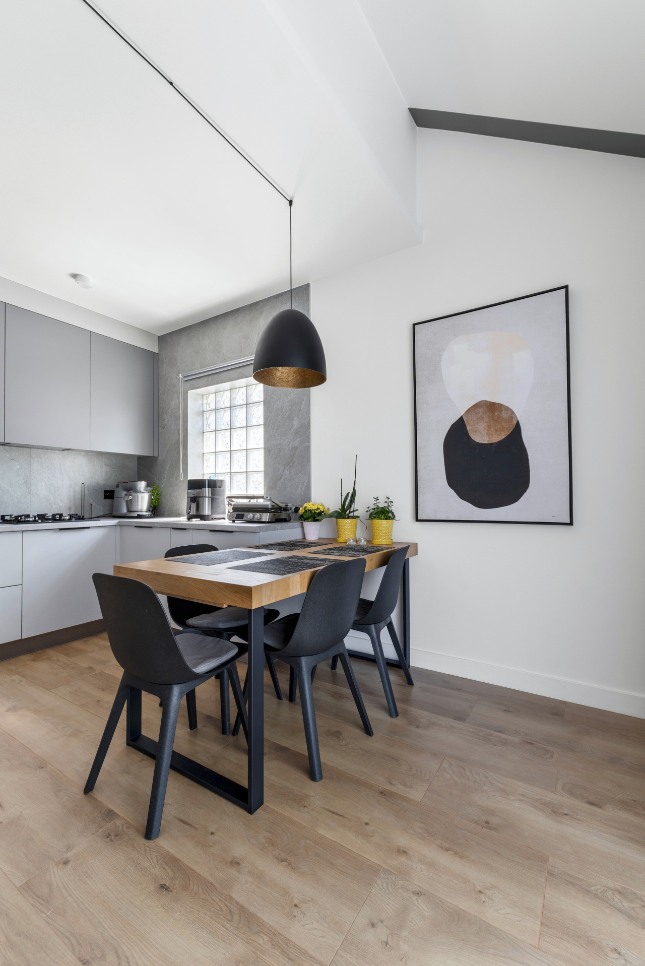 Nowoczesna kuchnia z szarymi szafkami i drewnianym stołem z czarnymi krzesłami. Nad stołem wisi czarna lampa. Na ścianie abstrakcyjny obraz w czarnej ramie.