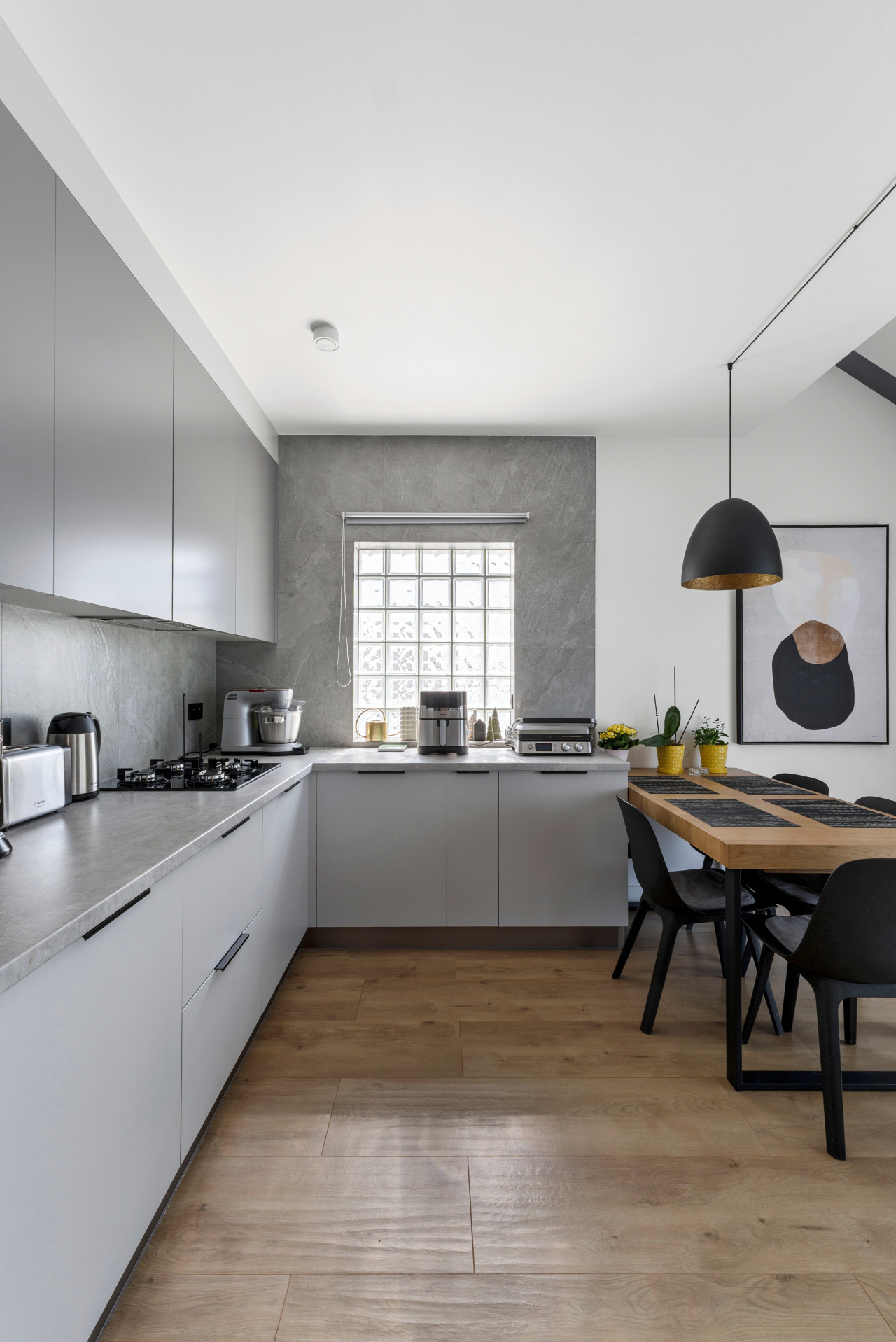 Nowoczesna kuchnia w szarościach z drewnianą podłogą i stołem jadalnym, oświetlona wiszącą lampą. Okno z luksferów w tle.