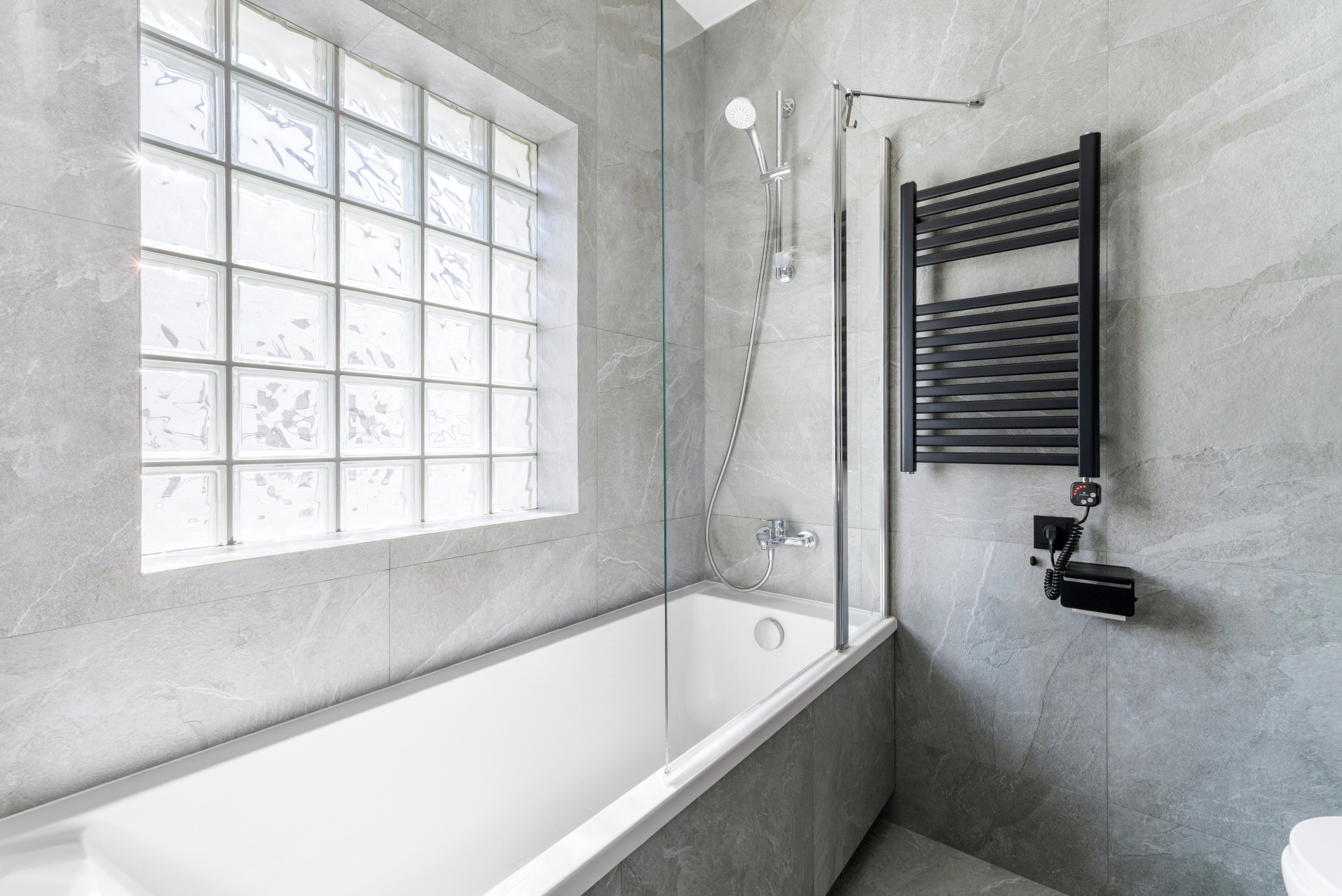 Szara łazienka z wanną, szybą prysznicową i oknem z luksferami. Czarny grzejnik drabinkowy na ścianie. Nowoczesny design, jasne wnętrze.