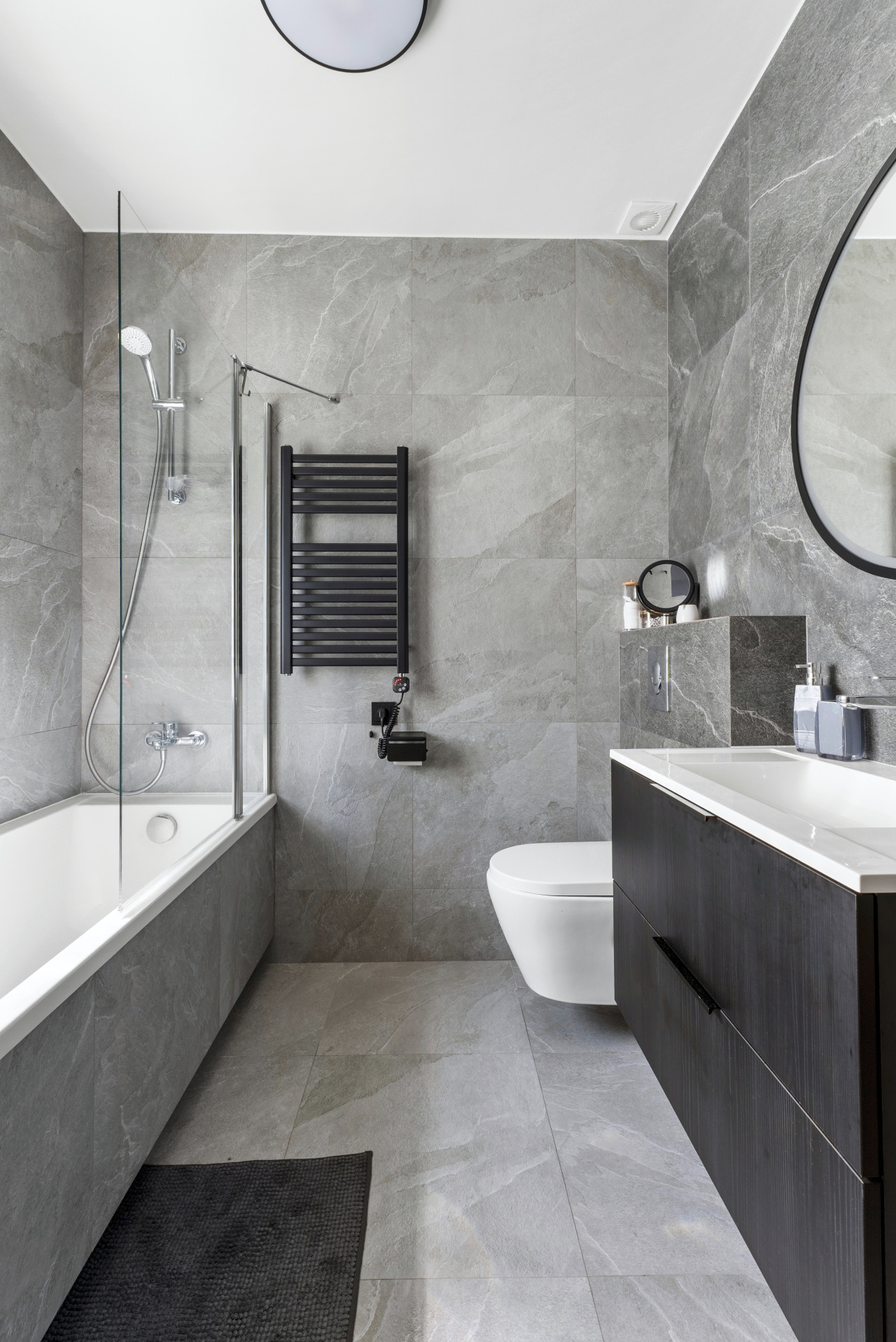 Szara łazienka z wanną, grzejnikiem drabinkowym i umywalką z szafką. Płytki imitujące kamień na ścianach i podłodze. Minimalistyczny design, okrągłe lustro.