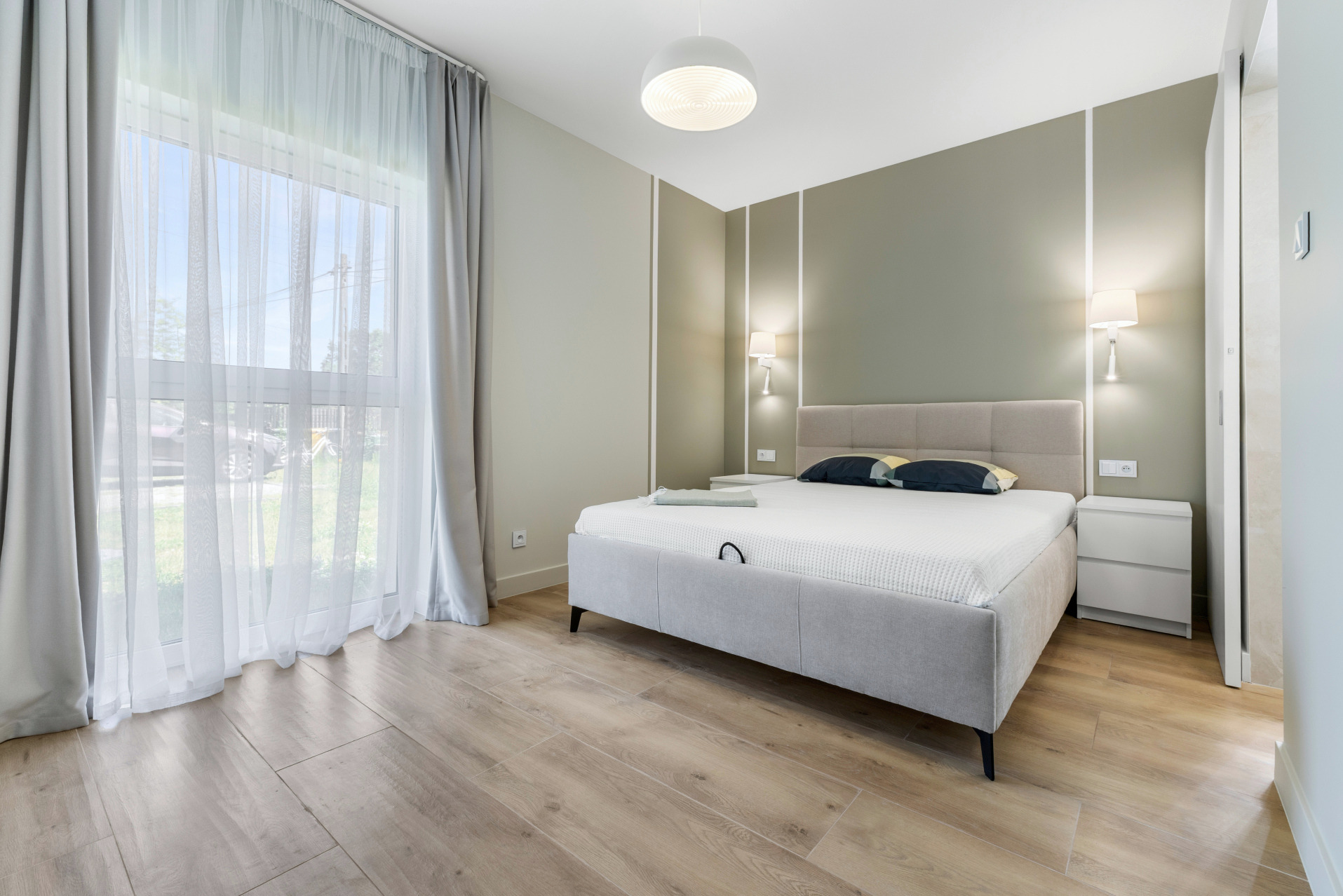 Jasna sypialnia w neutralnych barwach. Duże okno z zasłonami, łóżko z beżowym zagłówkiem i szafki nocne. Podłoga z paneli imitujących drewno. Lampy ścienne i sufitowa.