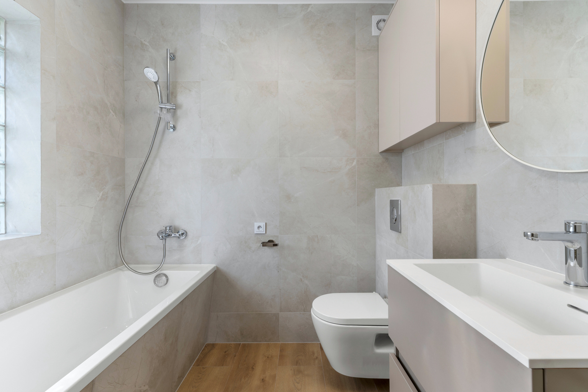 Jasna łazienka z beżowymi szafkami, wanną, umywalką i WC. Marmurowe płytki na ścianach i drewnopodobna podłoga. Okrągłe lustro.