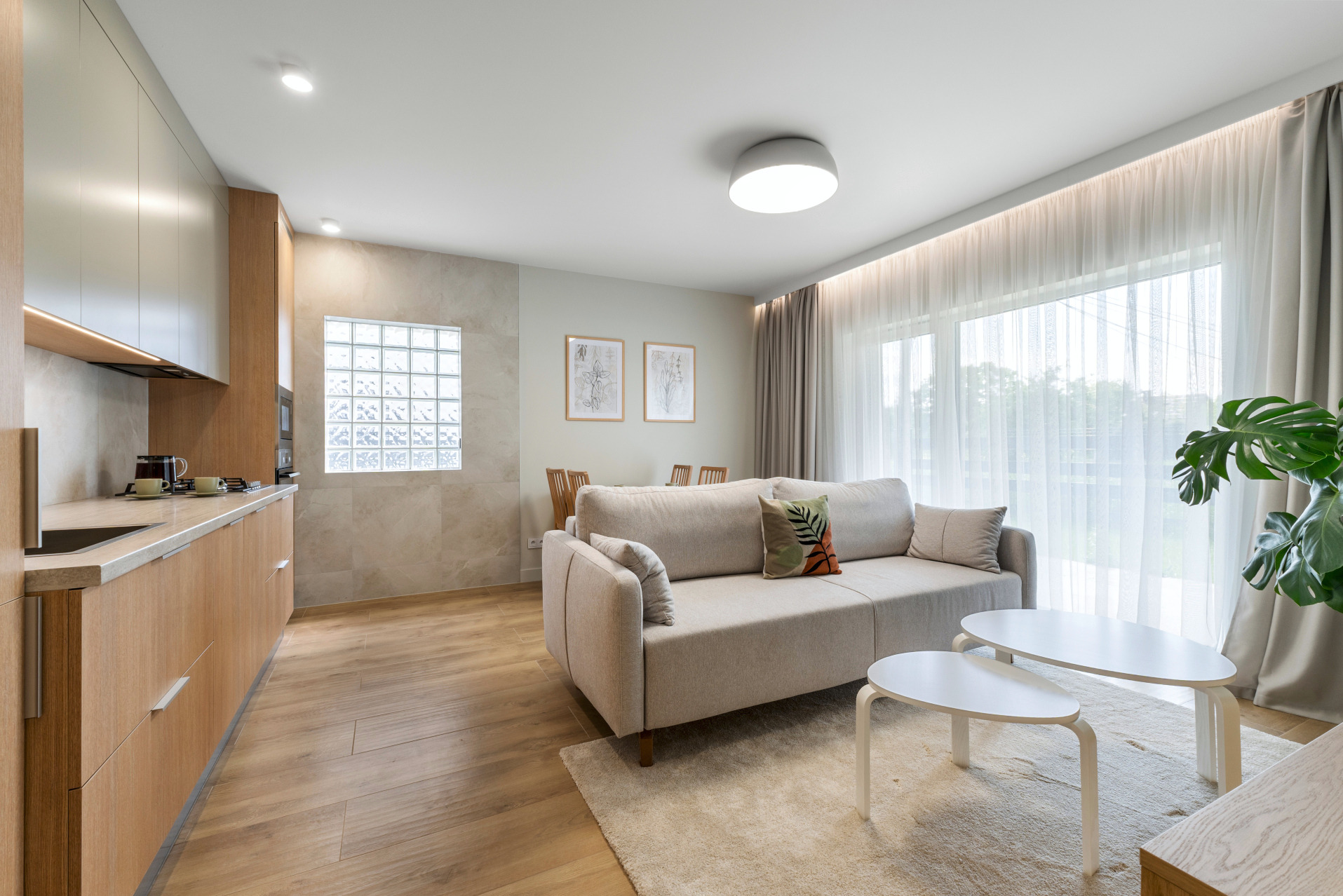 Jasne, nowoczesne wnętrze: drewniana podłoga, beżowa kanapa z poduszkami, białe stoliki kawowe, kuchnia z drewnianymi szafkami i okno z luksferami.