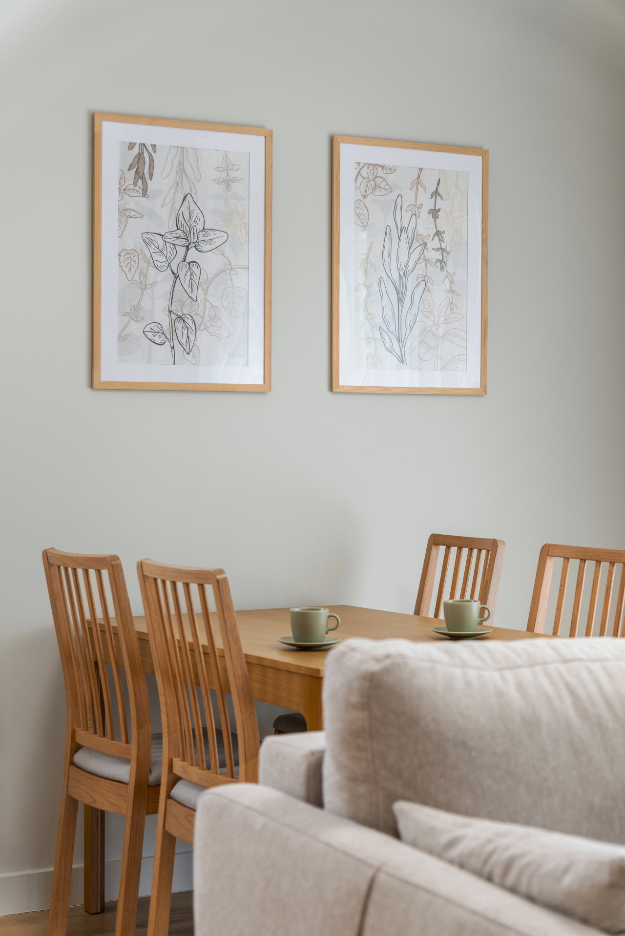 Jasne drewniane krzesła i stół z filiżankami na tle ściany z dwoma obrazami w drewnianych ramach. W tle widoczny fragment sofy.