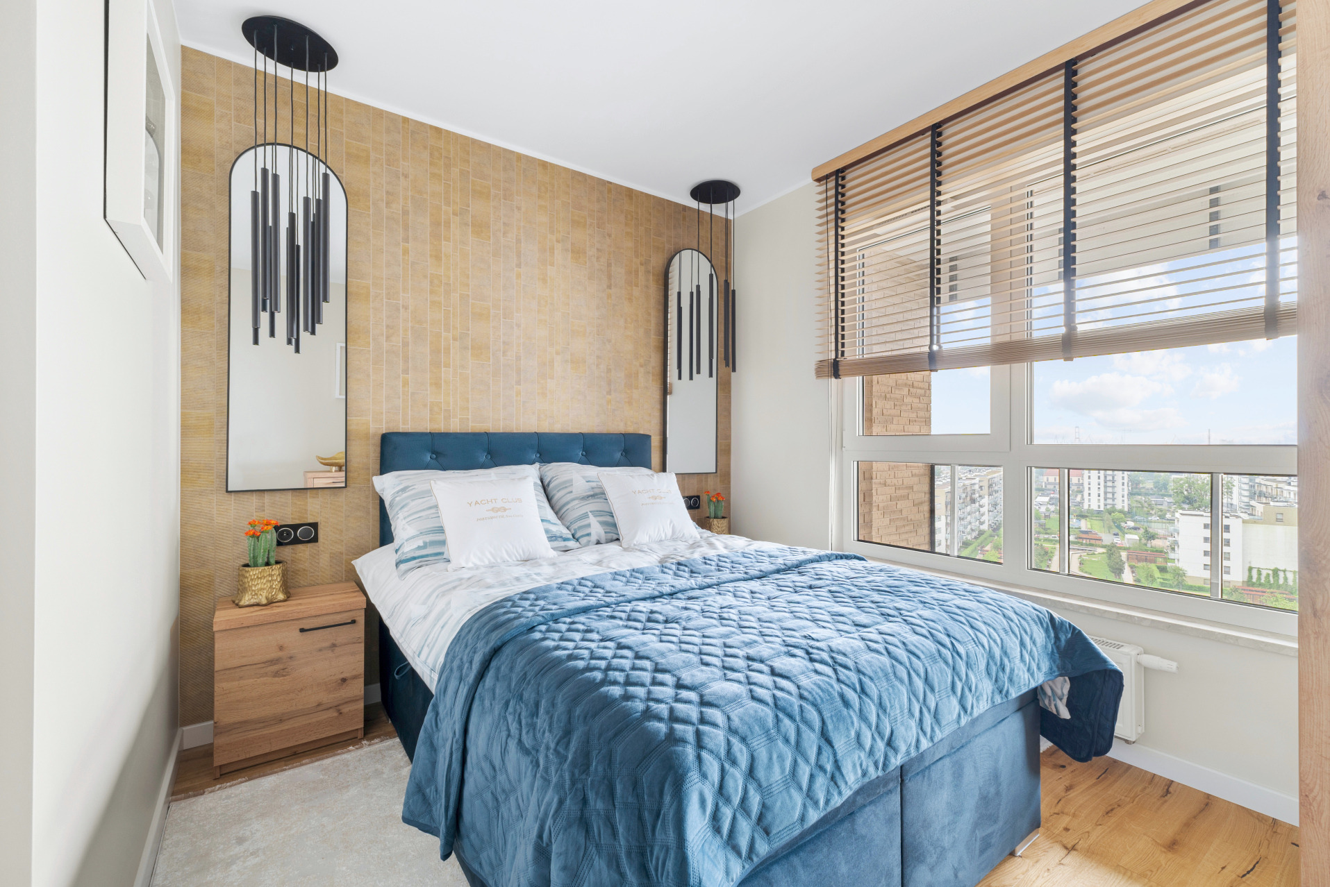 Sypialnia w odcieniach beżu i błękitu. Łóżko z pikowanym, błękitnym pledem, drewniana szafka nocna z kwiatami, okno z drewnianymi żaluzjami i lustra.