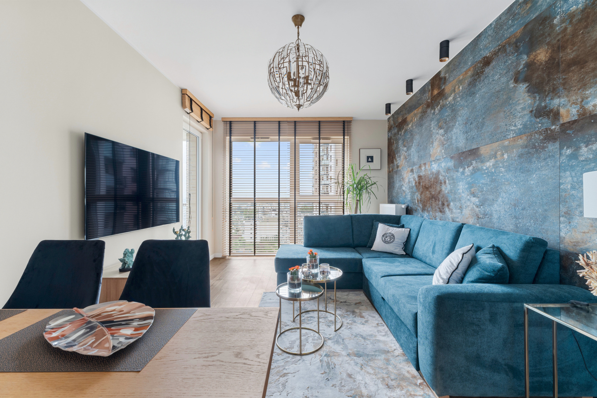 Stylowy salon z narożnikiem w odcieniach błękitu, dekoracyjną ścianą i okrągłym żyrandolem. Widok na miasto przez okno z żaluzjami.
