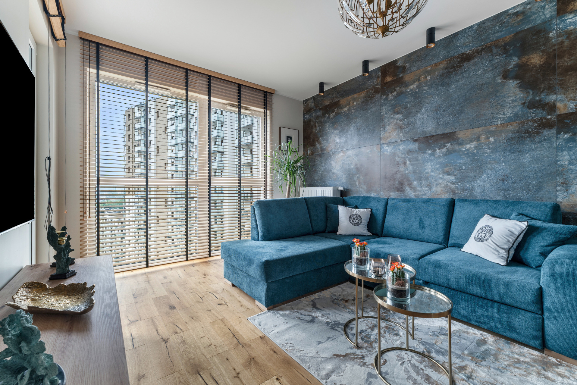 Stylowy salon z turkusową sofą narożną, stolikami kawowymi i zasłoniętymi żaluzjami oknami. Dekoracyjna ściana w odcieniach niebieskiego. Nowoczesne oświetlenie.