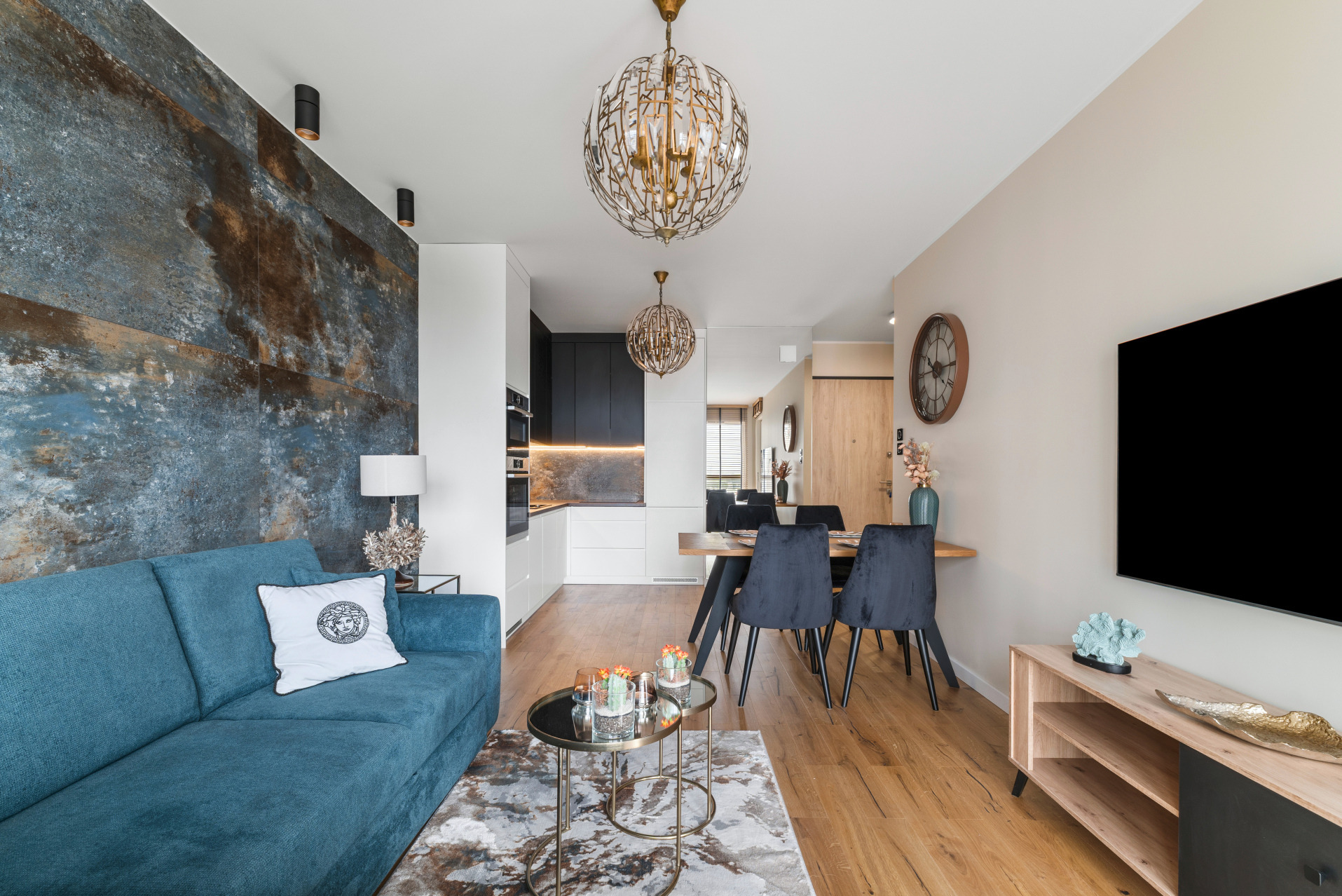 Nowoczesny salon z turkusową sofą, oryginalną ścianą i okrągłymi lampami. Widok na jadalnię i kuchnię. Parkiet na podłodze, stoliki kawowe i dekoracje.