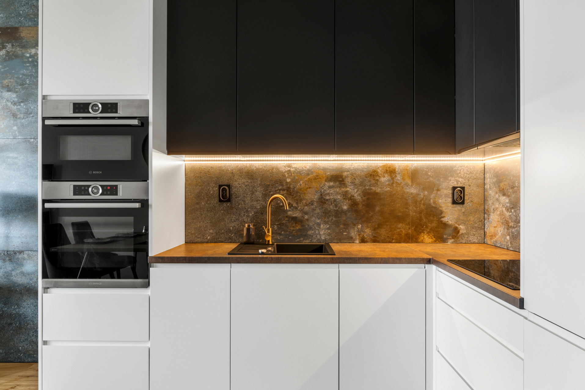 Nowoczesna kuchnia z białymi szafkami, czarnymi górnymi szafkami i złotą armaturą. Podświetlenie LED podkreśla unikalną fakturę ściany nad blatem.