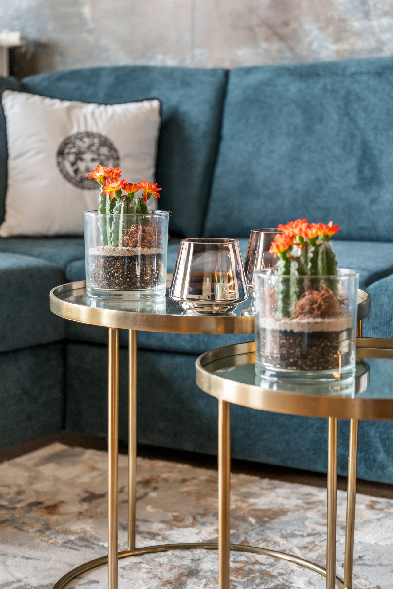 Dwa kaktusy z pomarańczowymi kwiatami w szklanych doniczkach na okrągłych, złotych stolikach. W tle turkusowa sofa i wzorzysty dywan.