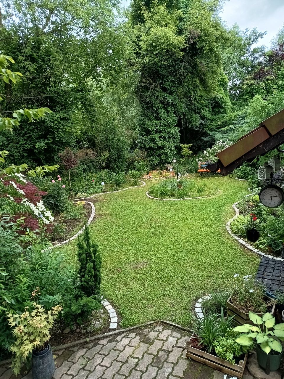 Zadbany trawnik z równo przystrzyżoną trawą, otoczony rabatami kwiatowymi i krzewami, widok z góry, z fragmentem zadaszenia i zegarem w tle.