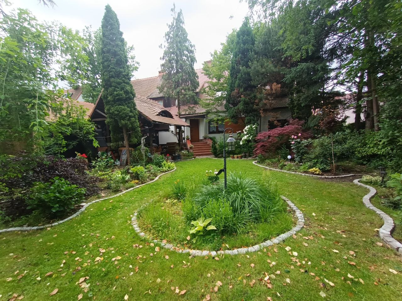 Urokliwy ogród przydomowy z okrągłą rabatą kwiatową i krętą ścieżką z kostki, prowadzącą do domu z czerwonym dachem. W tle bujna zieleń drzew.