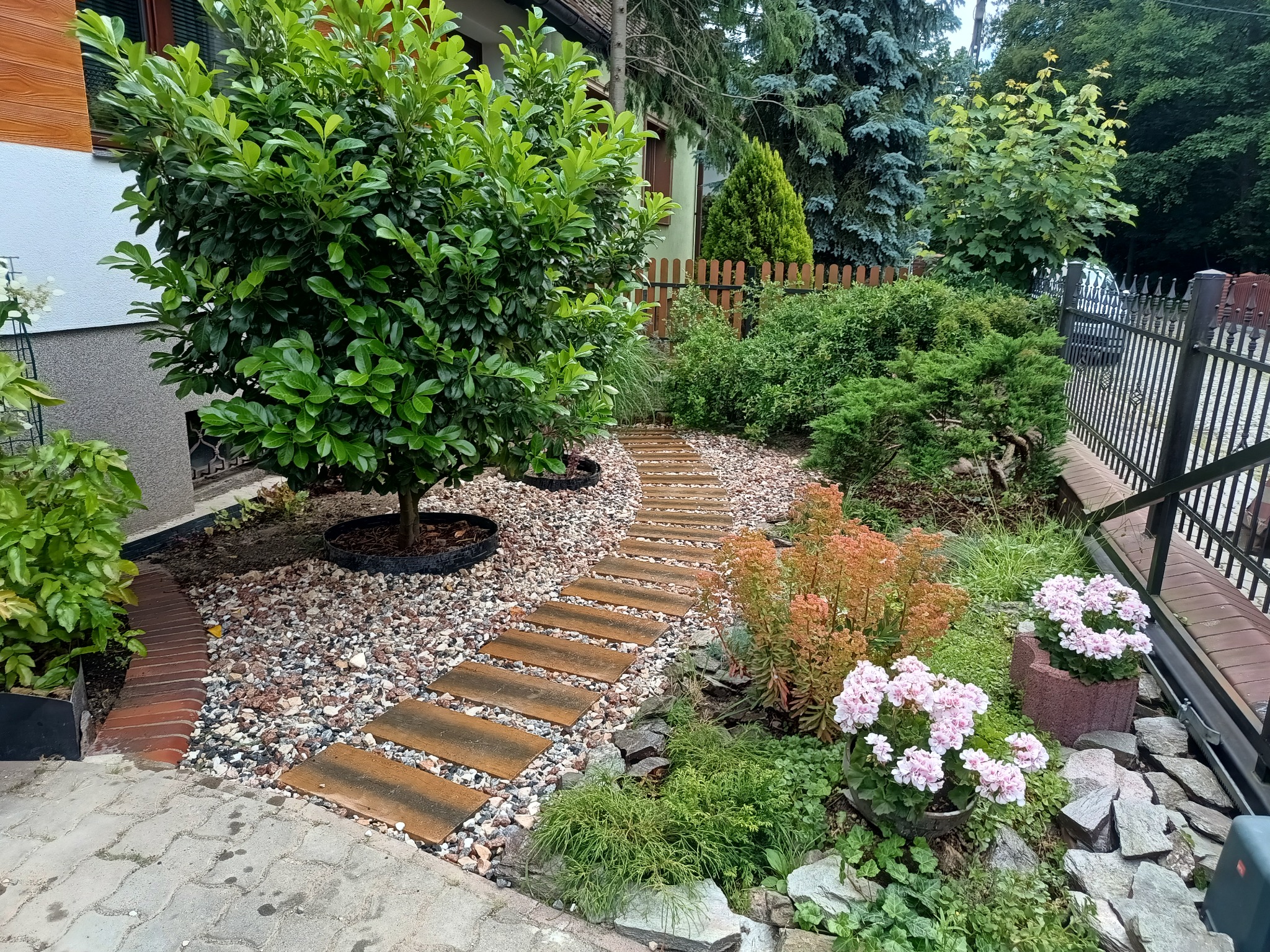 Ogród z ścieżką z płyt, obsadzony zielenią i kwiatami. Widoczne drzewa, krzewy, kamienie ozdobne. Ścieżka prowadzi wzdłuż domu i ogrodzenia.