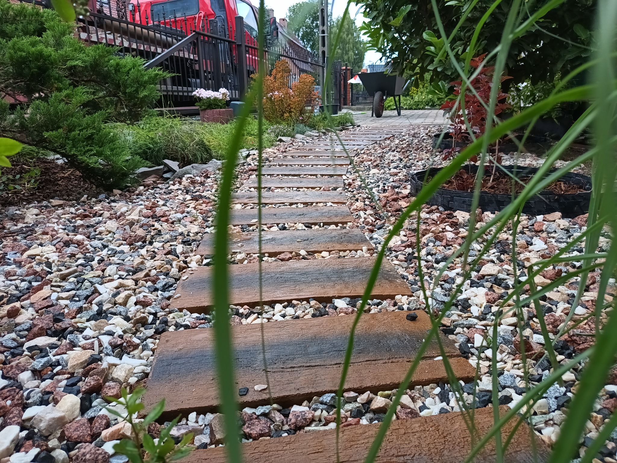 Ścieżka z drewnopodobnych płyt prowadząca przez ogród wysypany drobnymi kamieniami, z roślinami i taczką w tle. Ujęcie z niskiej perspektywy.