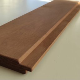 Artorix - Profilowana deska z drewna egzotycznego, widoczne frezy montażowe, oświetlona promieniami słonecznymi na gładkim tle. Detal wykończeniowy podłogi lub tarasu.