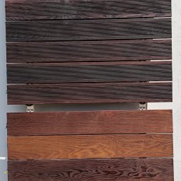 Artorix - Ekspozycja różnych odcieni desek tarasowych z drewna egzotycznego, zamontowanych na metalowych wspornikach na jasnym tle. Widoczne zróżnicowane faktury i kolory.