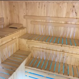 Artorix - Jasne drewno i niebieskie podświetlenie wewnątrz sauny. Wielopoziomowe ławy z szczelinami, pionowe deski na ścianach i suficie, ciepłe światło.