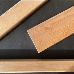 Artorix - Jasne, surowe deski z widocznym usłojeniem na czarnym tle. Jedna deska z kantem, pozostałe zaokrąglone. Detal materiału do wykończenia.