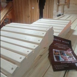 Artorix - Fragment konstrukcji z jasnego drewna z nawierconymi otworami, na tle drewnianej ściany i ulotki firmy 'Green Wood' z adresem i numerem telefonu.