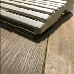 Biały, drewniany podnóżek z zaokrąglonymi szczebelkami, umieszczony na podłodze z paneli imitujących naturalne drewno. Widoczne łączenia i faktura drewna.