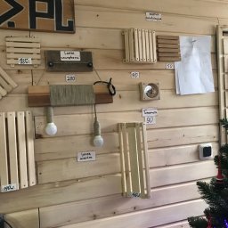 Artorix - Ekspozycja drewnianych elementów: kratki wentylacyjne, lampy i termometr na ścianie z boazerii. Widoczne ceny produktów i częściowo choinka.