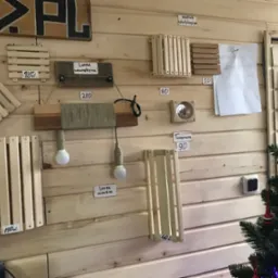 Ekspozycja drewnianych elementów: kratki wentylacyjne, lampy i termometr na ścianie z boazerii. Widoczne ceny produktów i częściowo choinka.