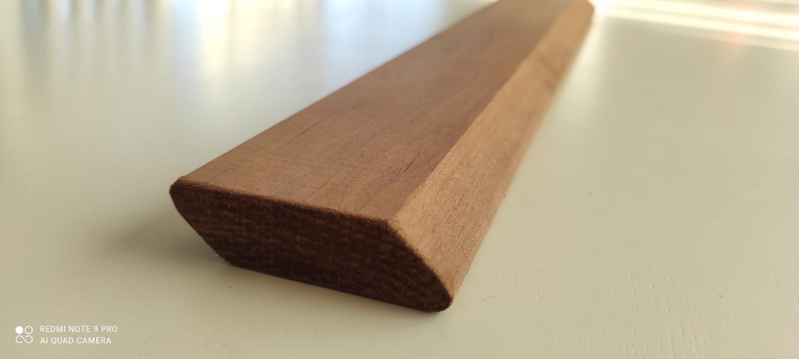 Fragment obrobionej deski z wyraźnym usłojeniem, leżącej na jasnej powierzchni. Kształt trapezu nadaje unikalny charakter. Widoczne detale cięcia i wykończenia.
