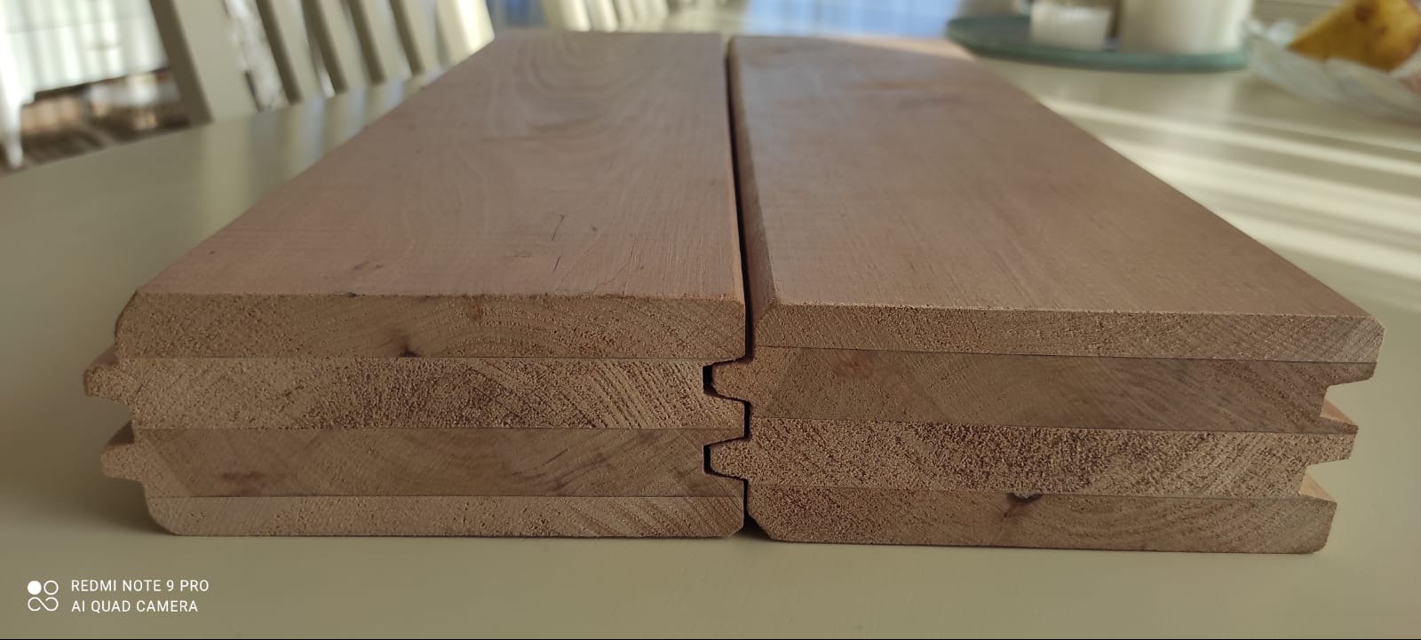 Profilowane deski z jasnego drewna, ułożone warstwami, ukazujące system łączenia pióro-wpust. Widoczne słoje drewna i precyzyjne wykończenie krawędzi.